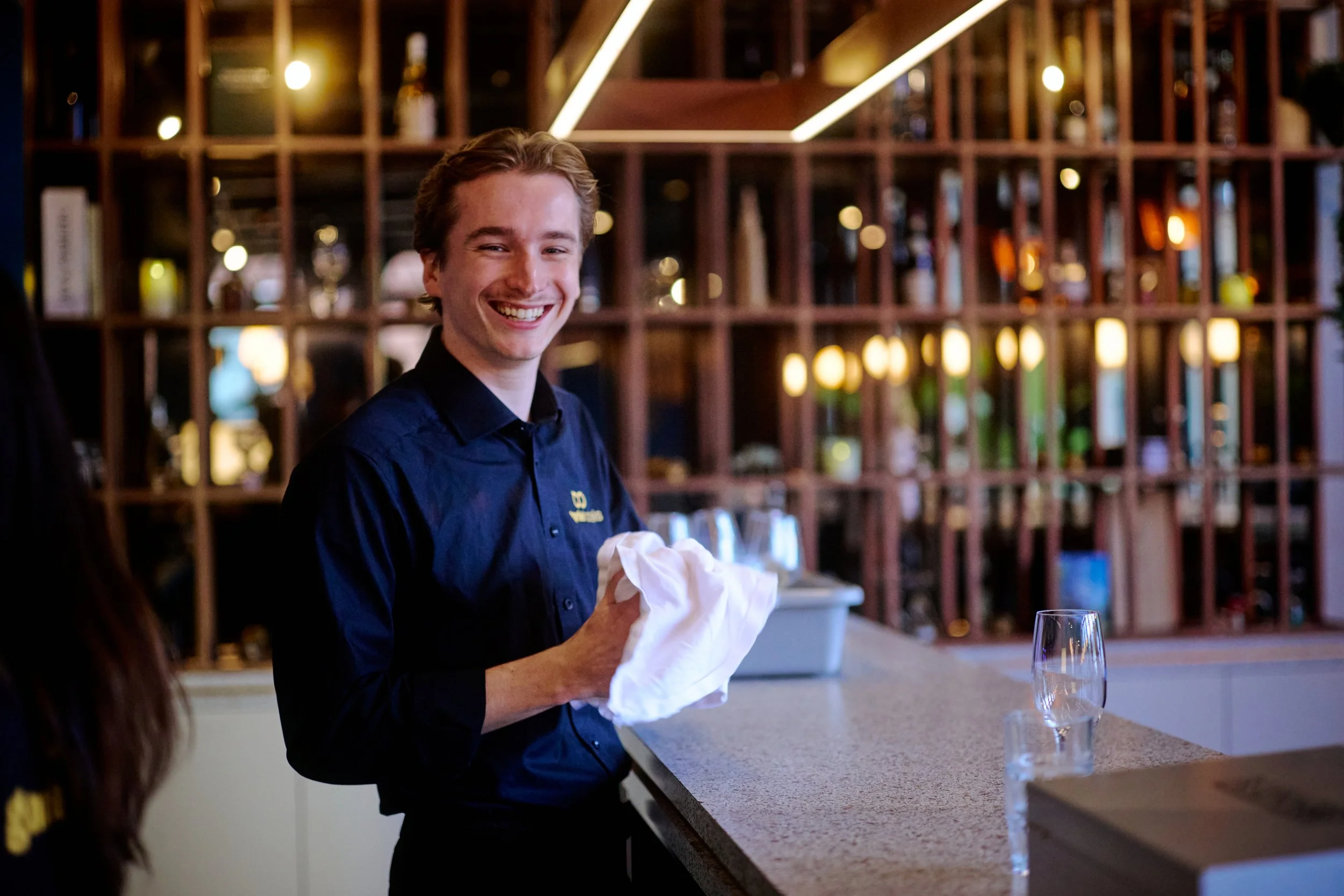 Smiling young man in a dark shirt, likely a waiter, standing at a bar or counter in a dimly lit restaurant or bar, holding a white cloth.