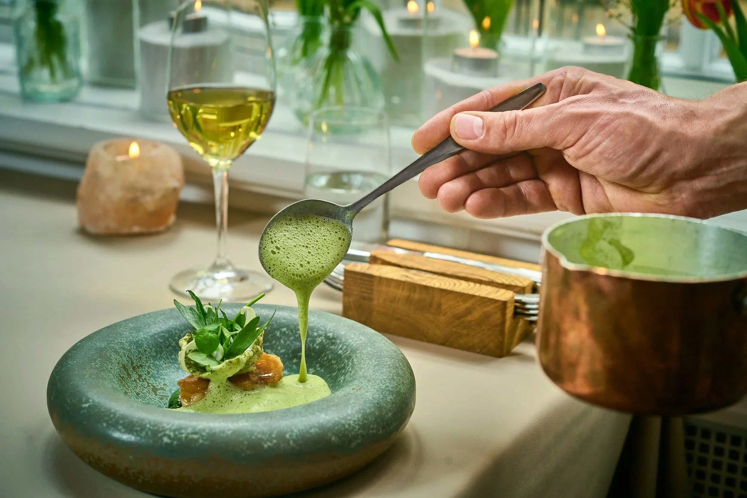 A plated dish with green sauce, garnished with herbs, with a hand pouring additional green sauce from a copper pitcher, set on a table with a wine glass, candles, and a blurred background of window and plants.