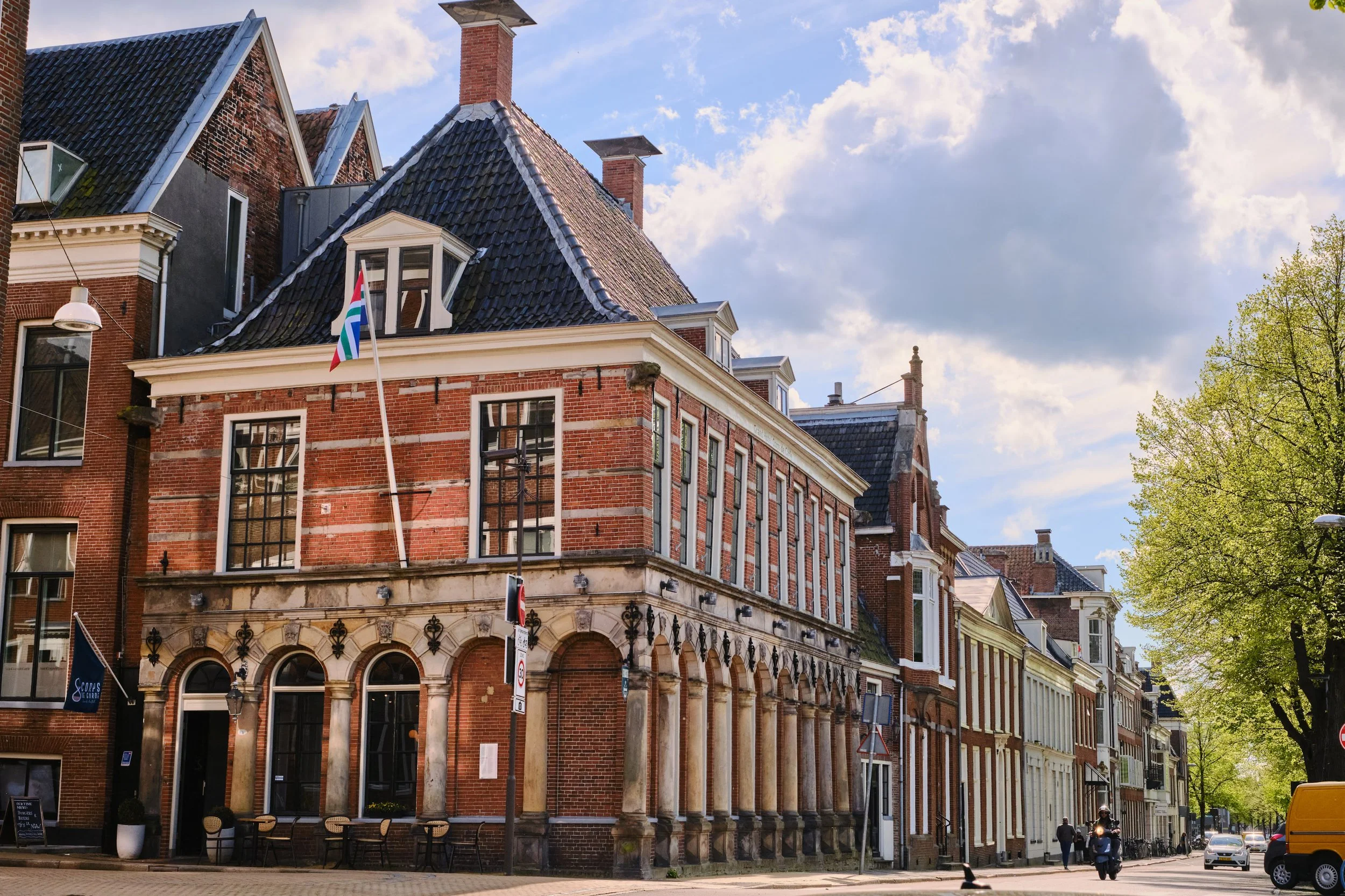 Street view of historic brick buildings with large windows, arched entryways, and outdoor seating in front, under a partly cloudy sky with some trees on the right side.