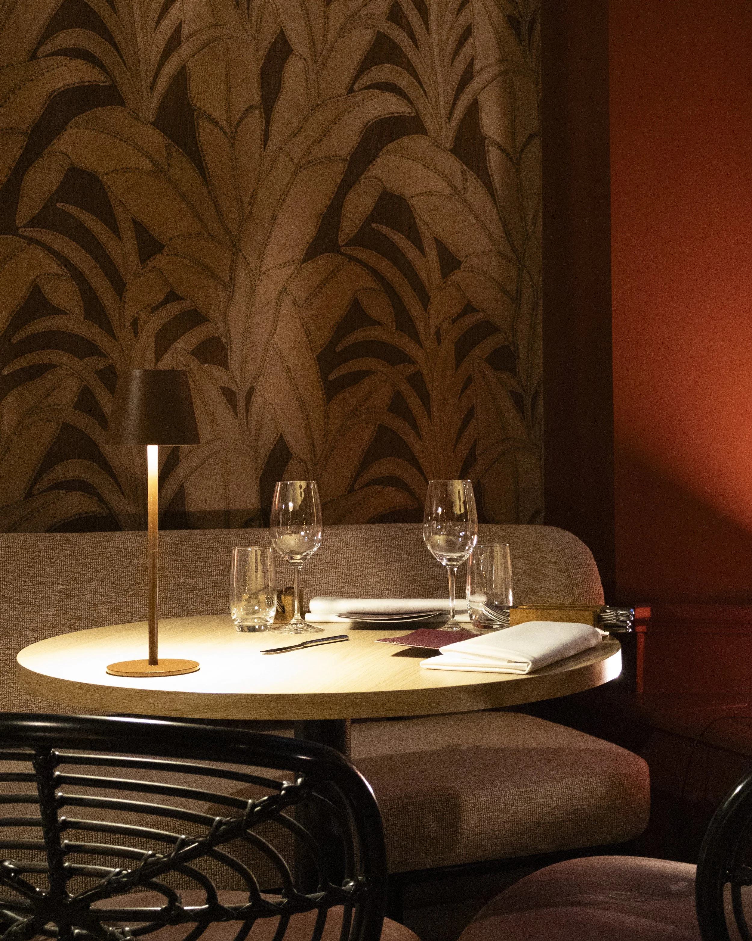 A restaurant table set with wine glasses, cloth napkins, and cutlery, illuminated by a warm table lamp. The background features a decorative wallpaper with a large leaf pattern and warm-colored walls.