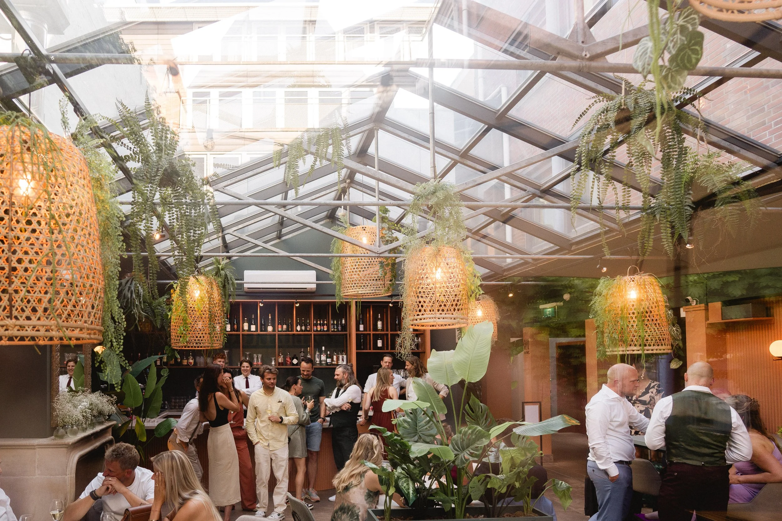 A lively scene inside a glass-roofed restaurant or café with hanging wicker light fixtures, lush green plants, and a crowd of people socializing and dining near a bar area.