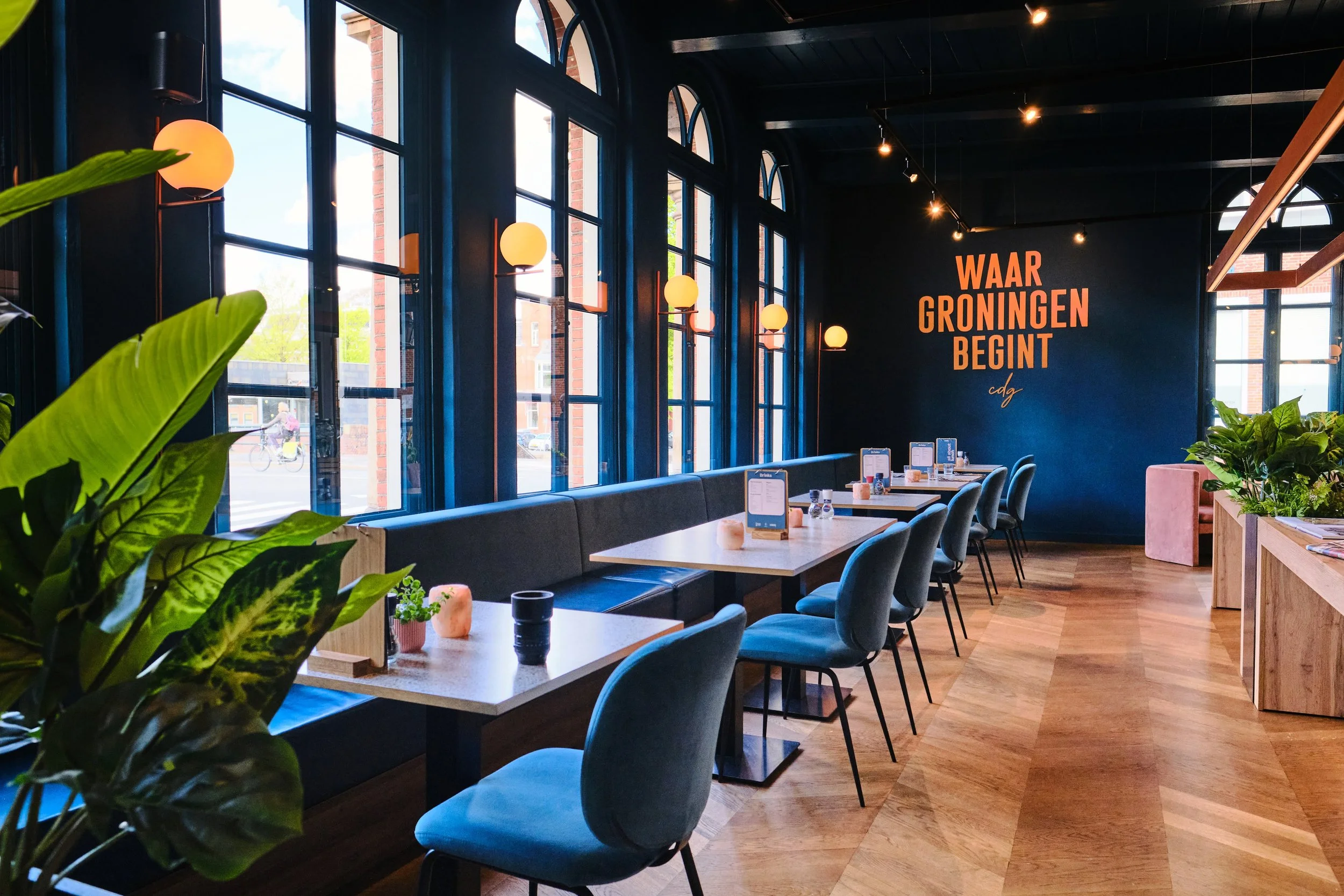 Interior of a modern restaurant or cafe with large arched windows, dark blue walls, and pendant lights. There are tables with menus, chairs, plants, and a wall with a sign reading 'WAAR GRONINGEN BEGINT'.