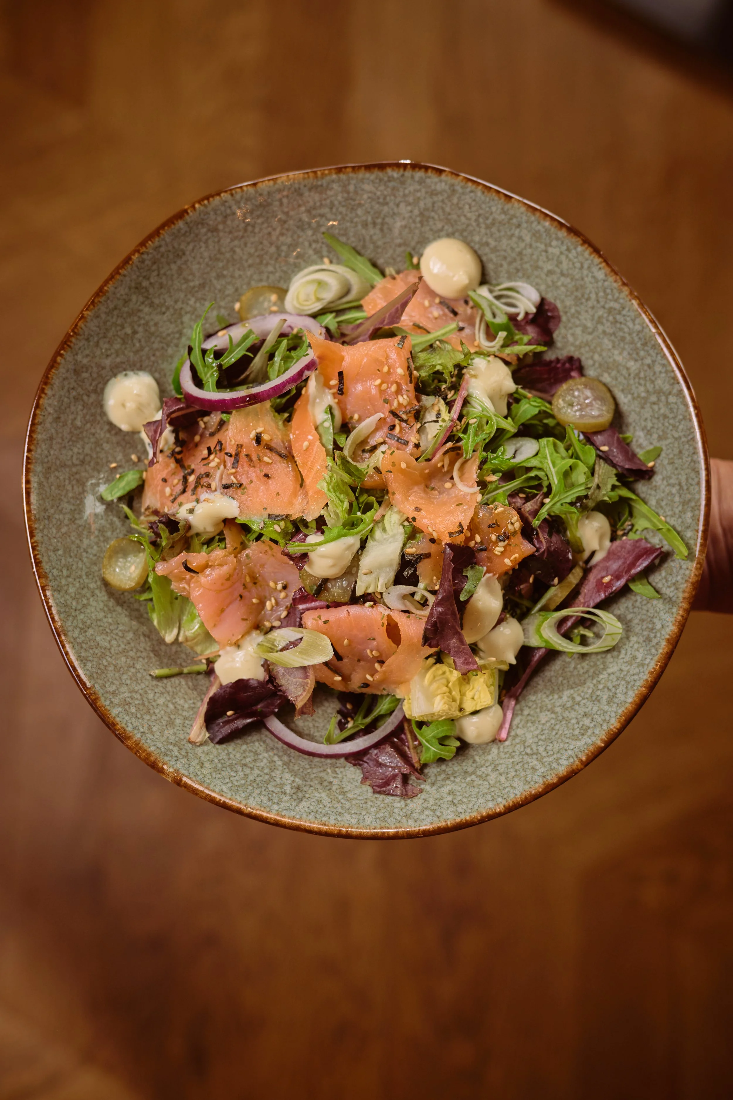 A bowl of mixed green and purple leafy salad with sliced onions, smoked salmon, creamy dressing, sesame seeds, and capers on a wooden table.