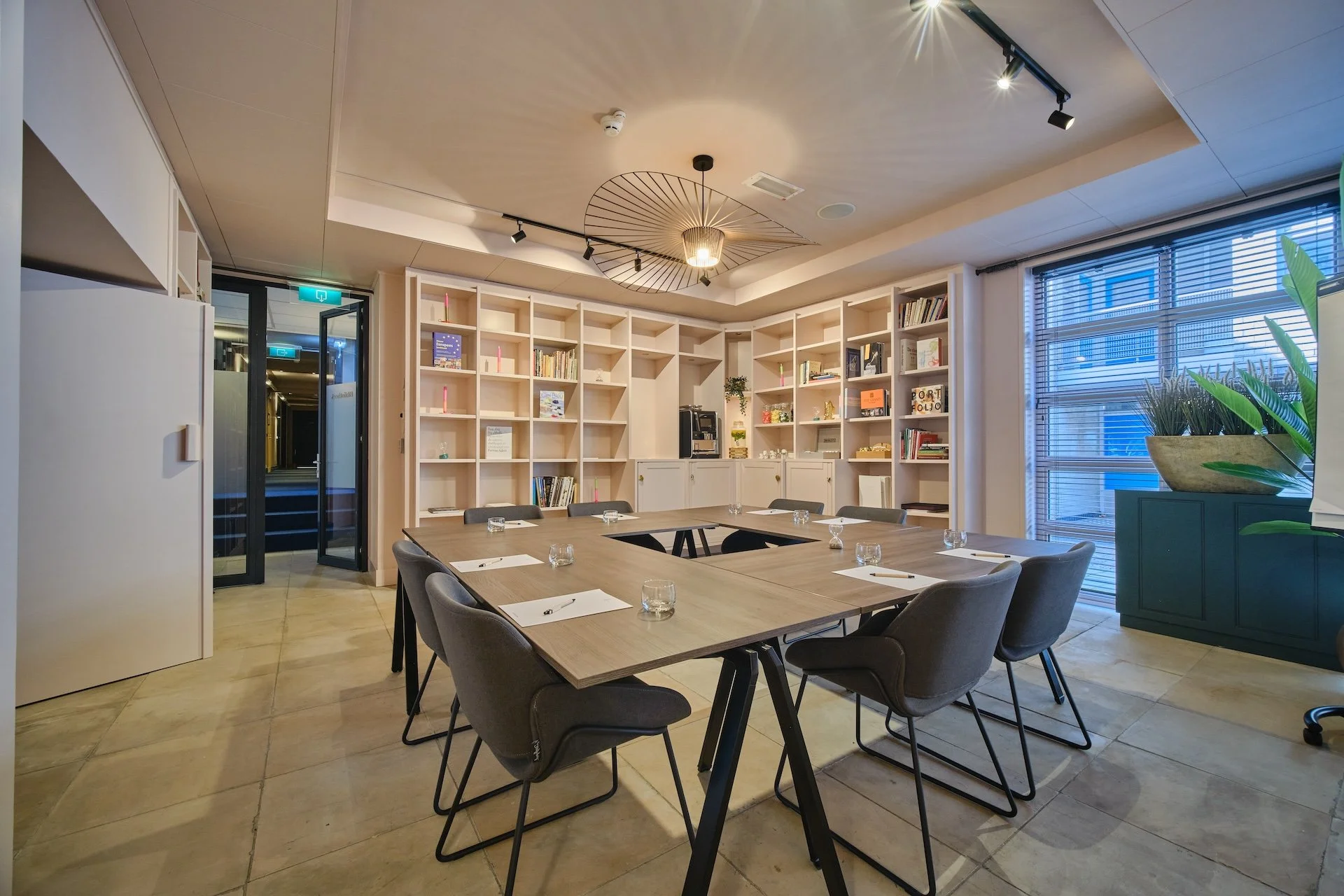 A modern conference room with a long wooden table, eight chairs, a large window with blinds, a bookshelf, ceiling lighting, and a decorative ceiling fixture.