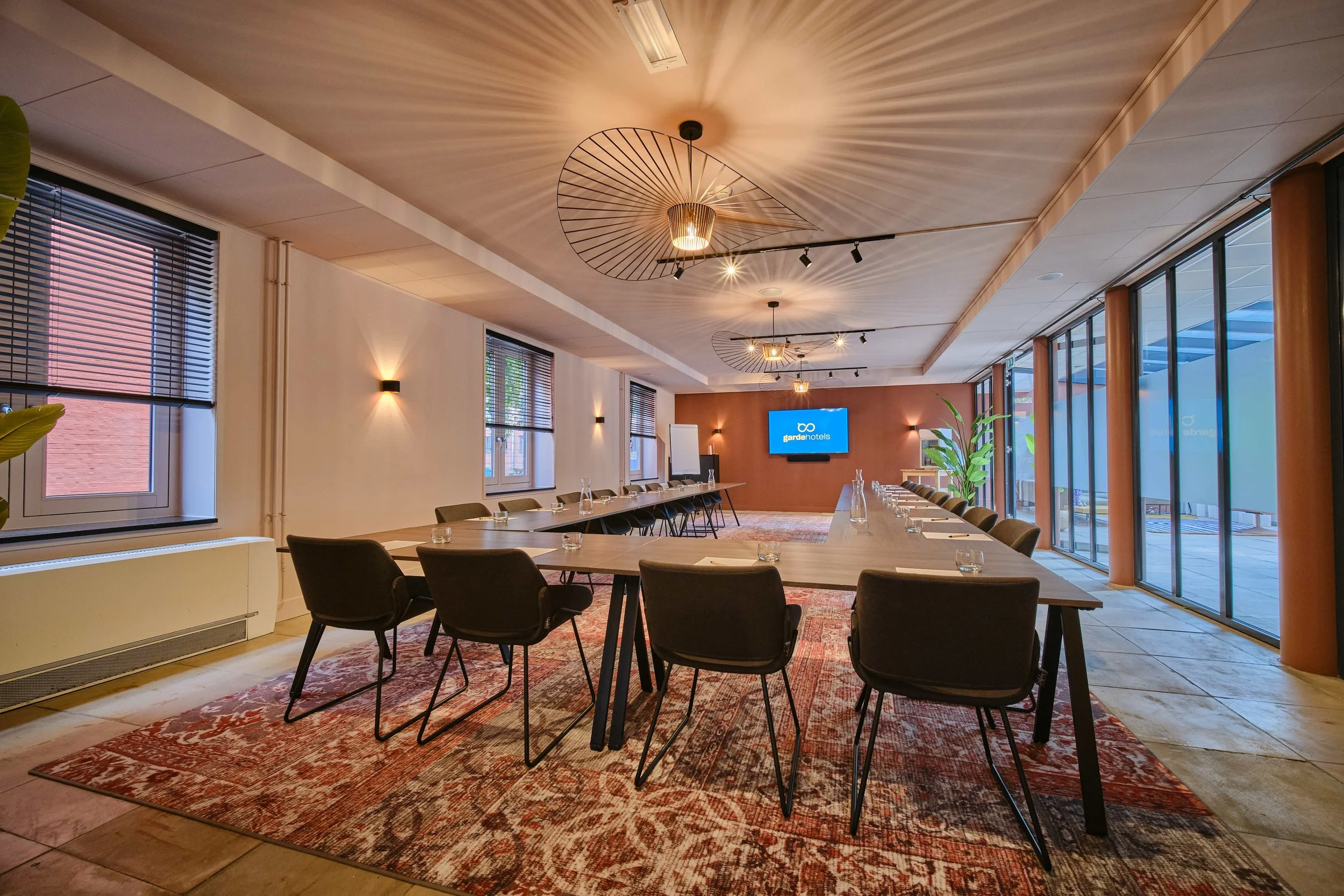 A modern conference room with a long rectangular table surrounded by black chairs, set with water glasses and papers. The room features large windows with blinds, pendant lighting fixtures on the ceiling, a flat-screen TV displaying 'gardehotels,' an