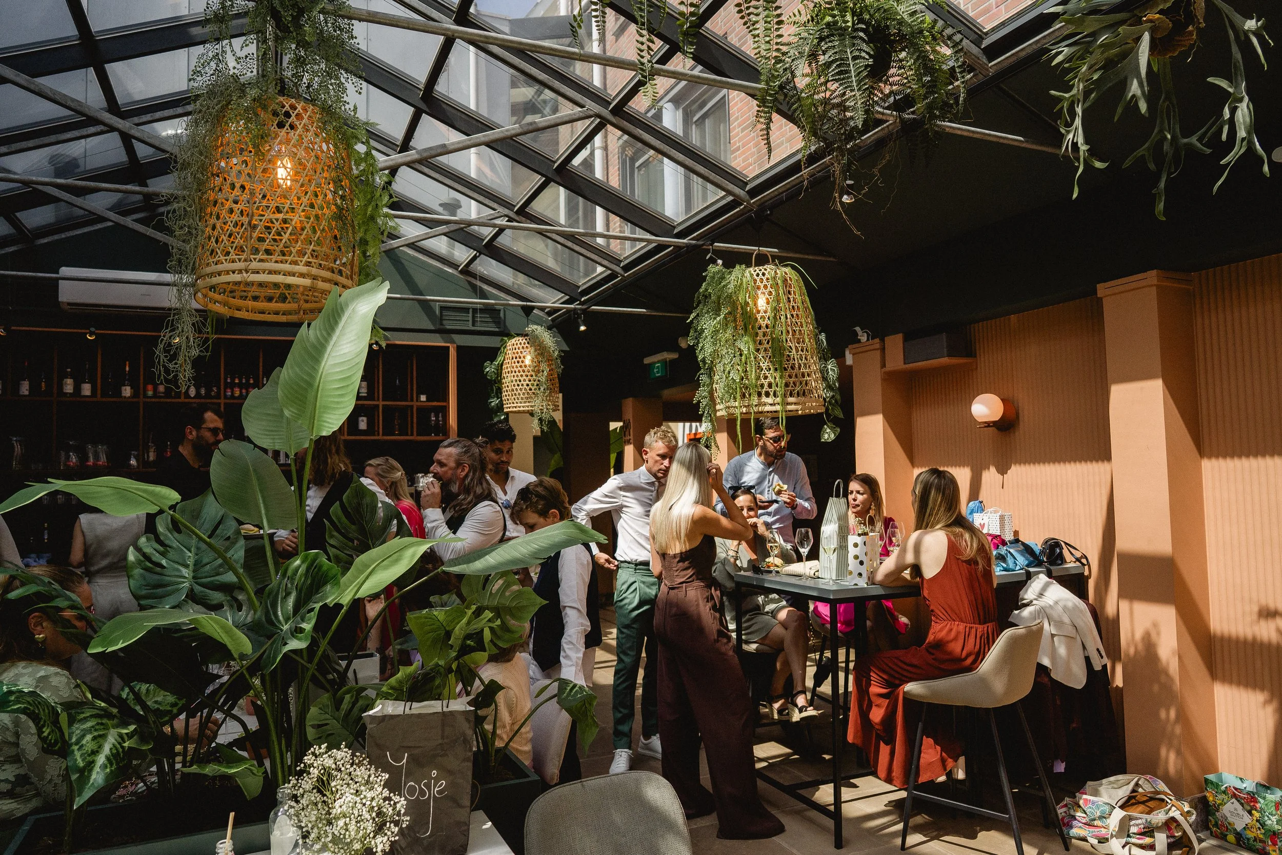 People gather in a modern indoor space with large glass ceiling windows, hanging wicker light fixtures, and lush green plants, engaging in conversations and enjoying an event.