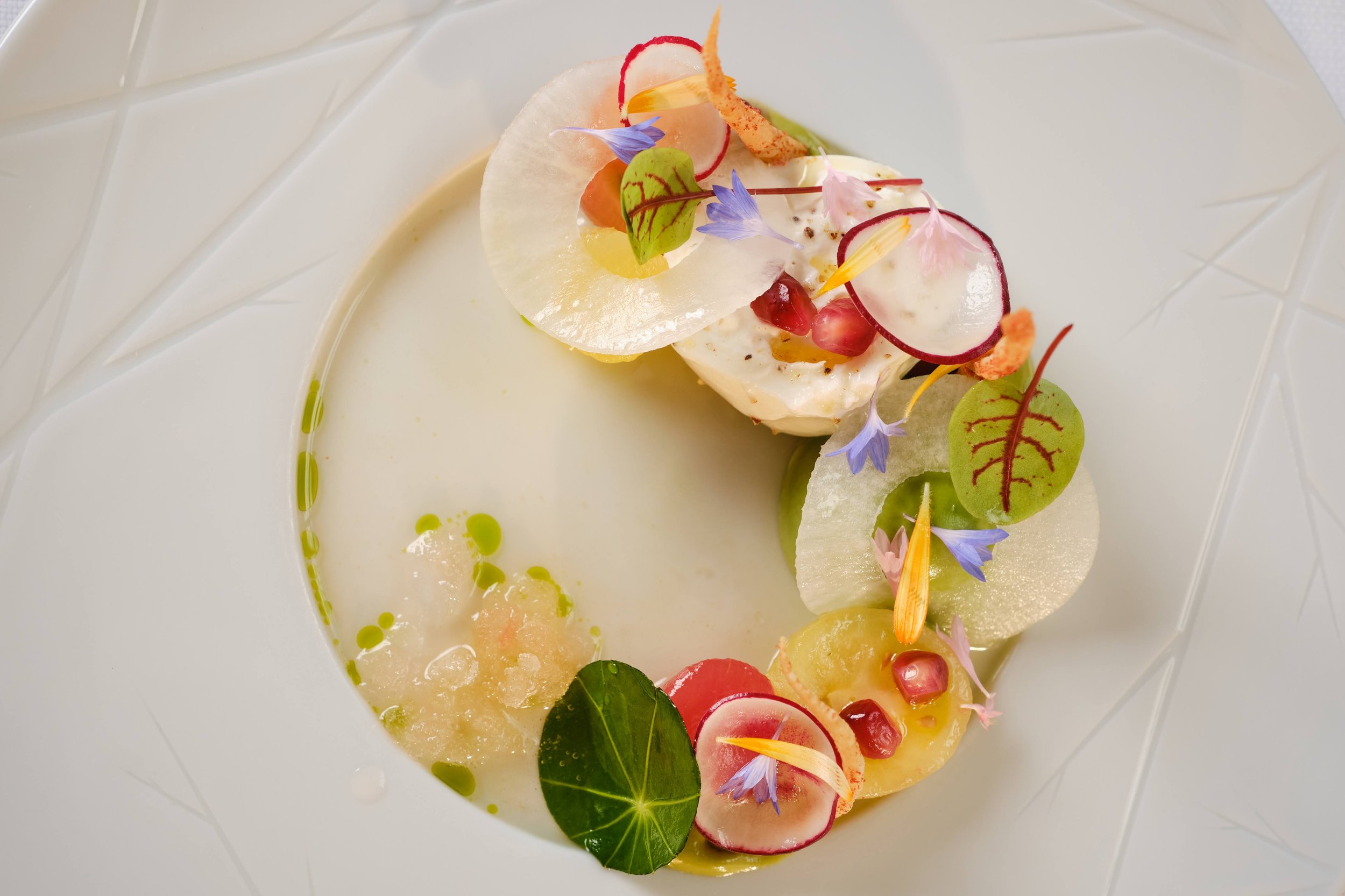 A plated dish of gourmet food with slices of radish, thinly sliced fruit or vegetable, edible flowers, microgreens, and green oil or sauce on a white plate.