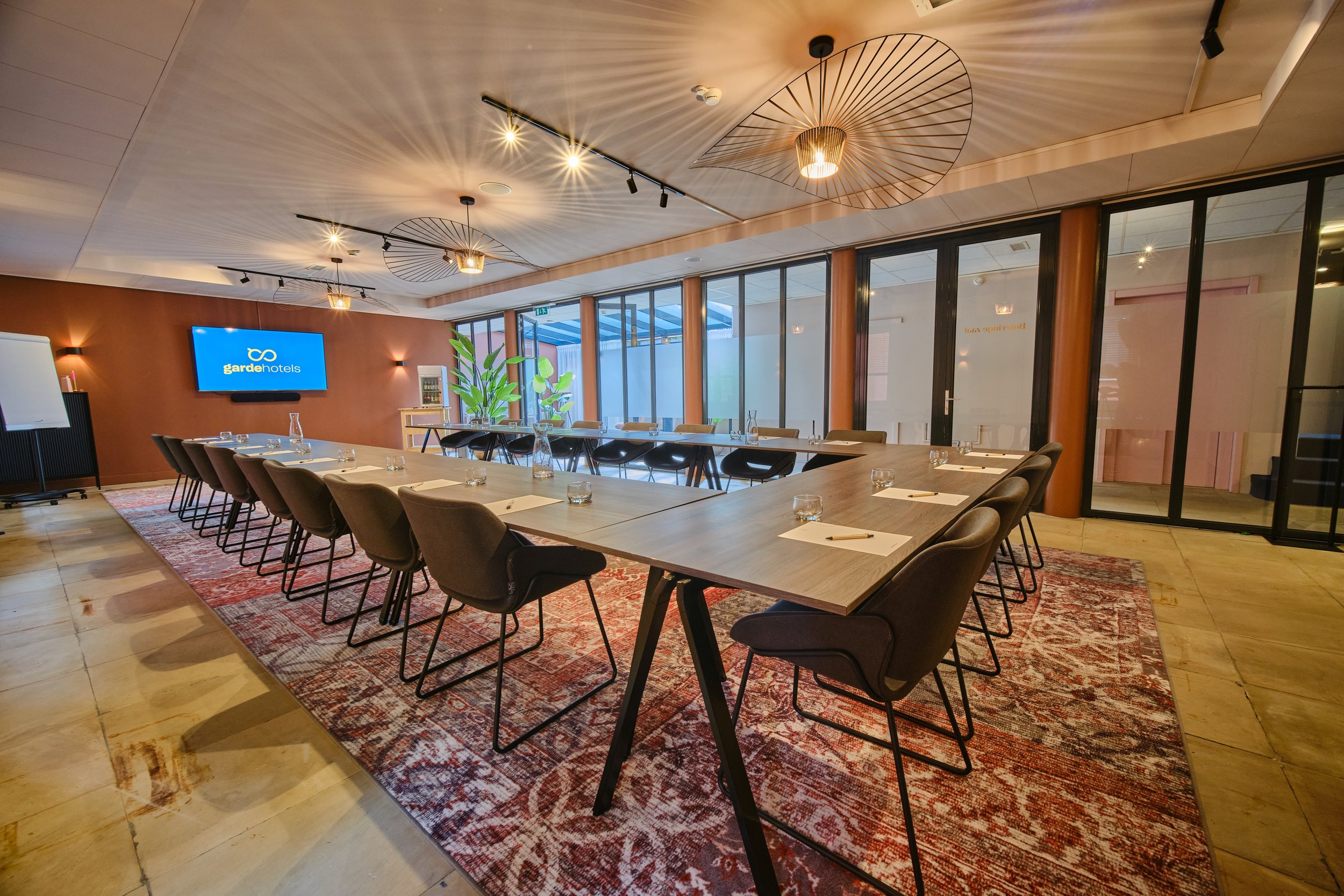 A modern conference room with a long wooden table, black chairs, and a patterned red rug on a light wooden floor. The ceiling has decorative lighting fixtures and wooden sunburst designs. A large flat-screen TV displaying the Garde Hotels logo is mounted on a maroon accent wall. Large windows with black frames let in natural light, and a potted plant is positioned near the window.