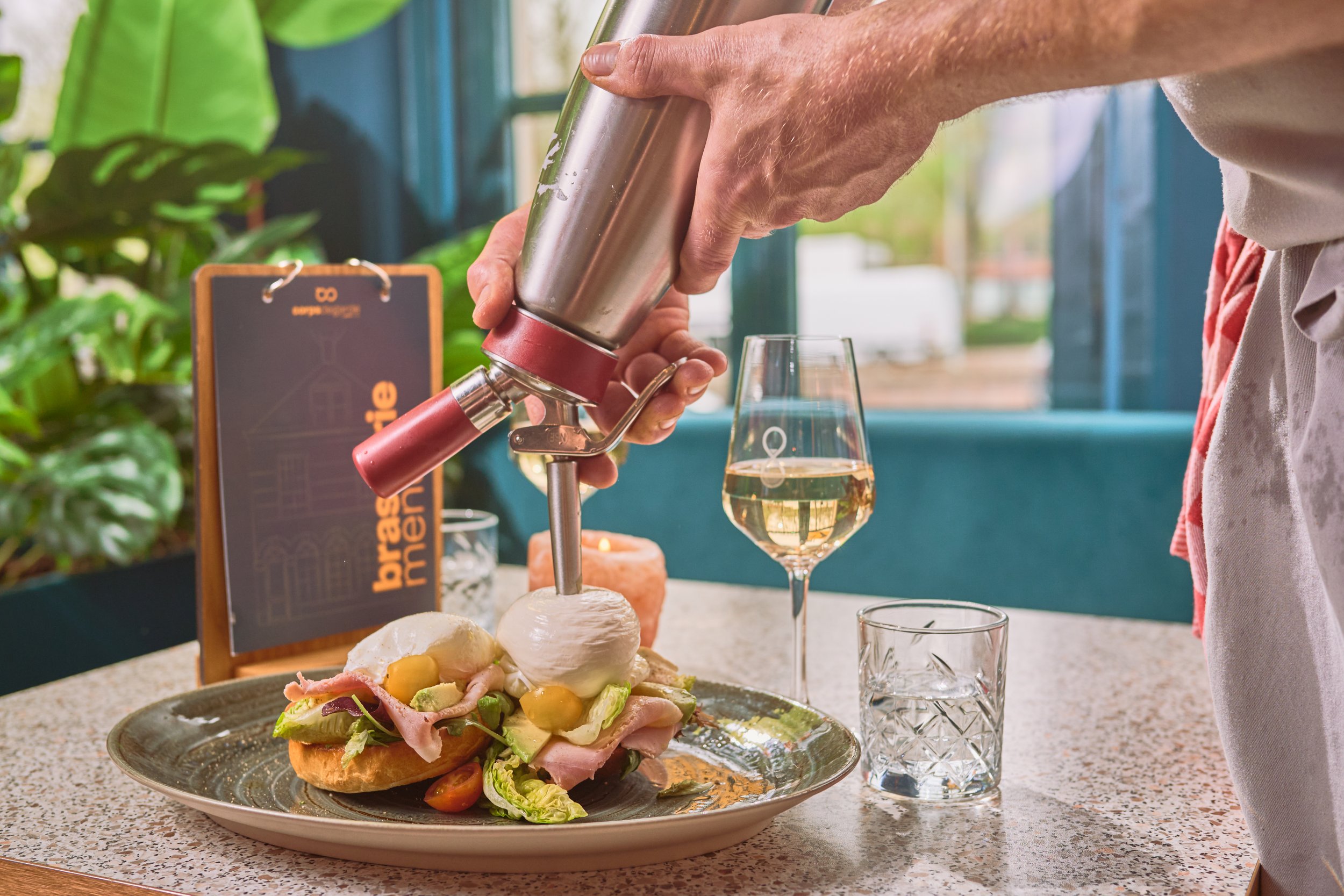 Person pouring liquid into a glass at a restaurant table with a salad on a plate.
