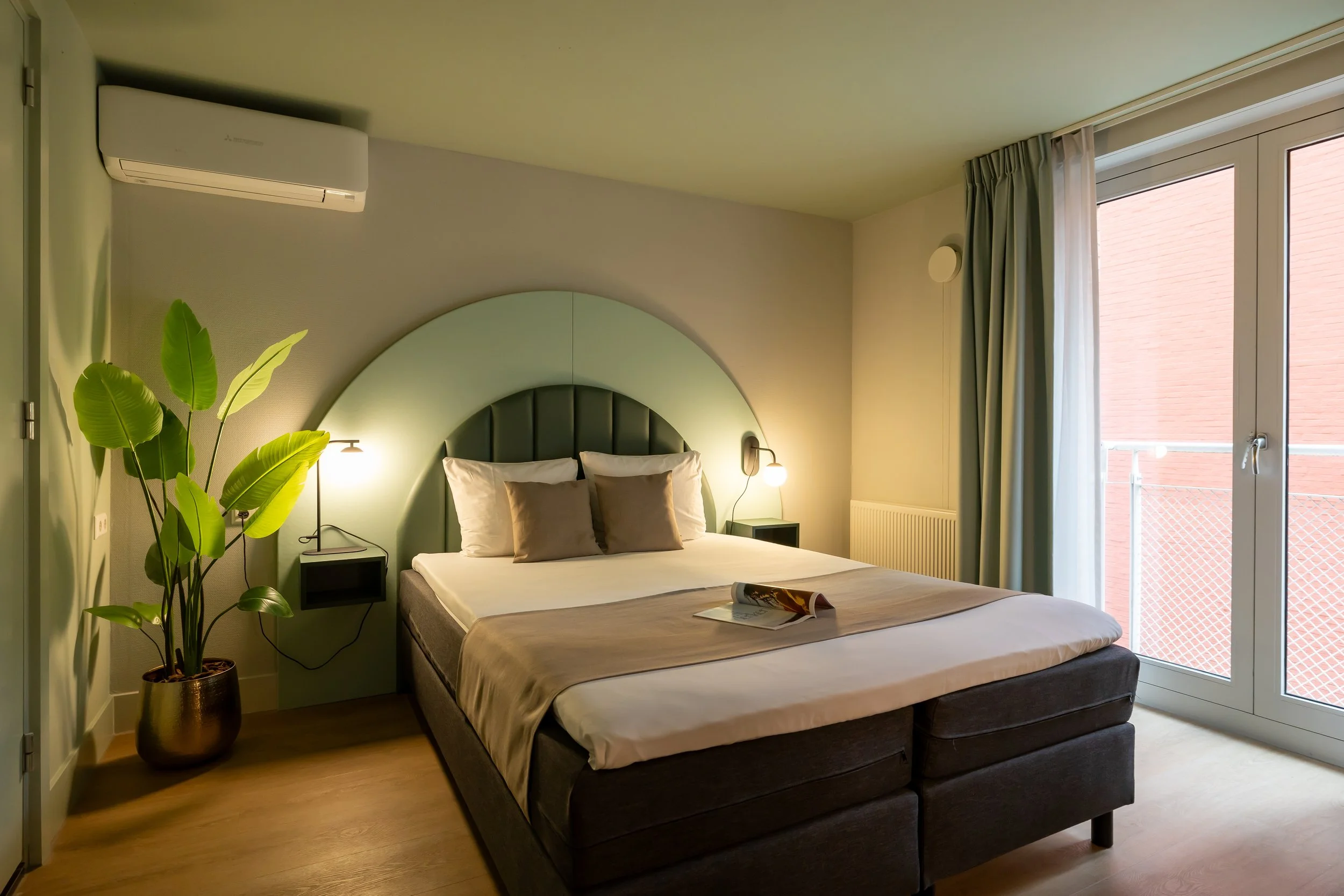 Modern bedroom with a large bed, gray headboard, potted plant with large green leaves, wall-mounted lighting, and a large window with curtains showing a brick building outside.