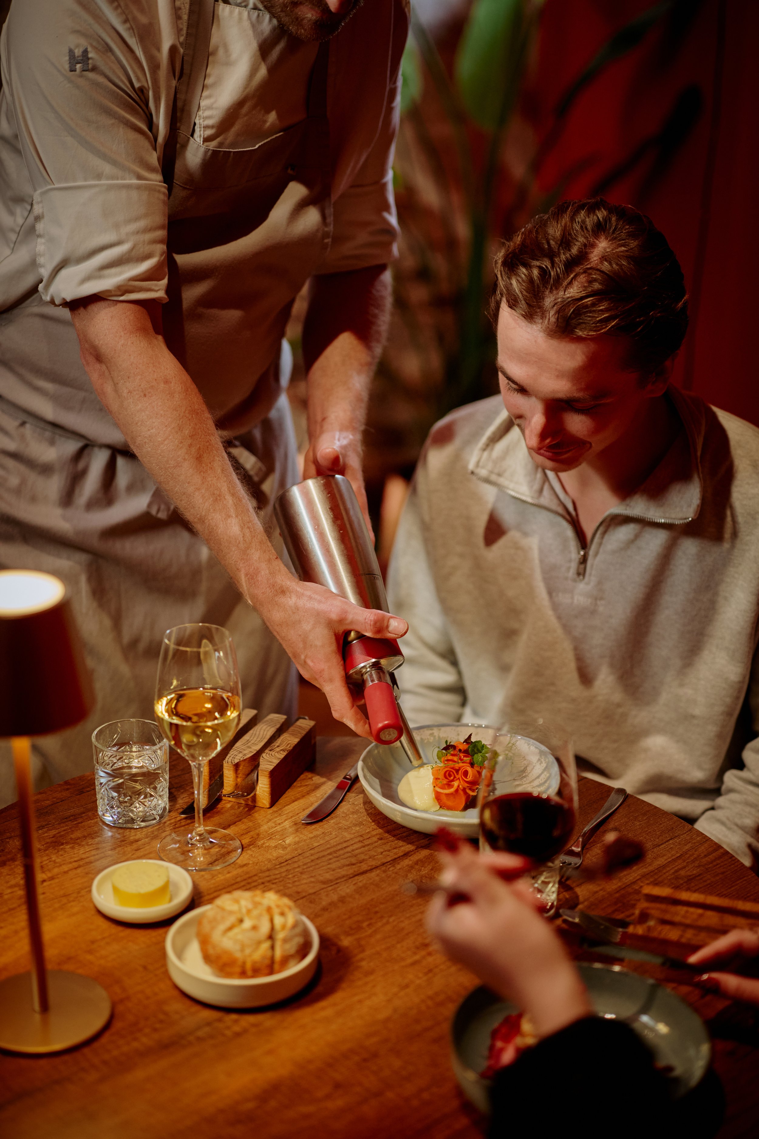 A person in a beige jacket is pouring a sauce or dressing over a plated dish of salmon, potatoes, and garnishes at a wooden table set with wine glasses, lemon slices, and small dishes, in a warmly lit restaurant.