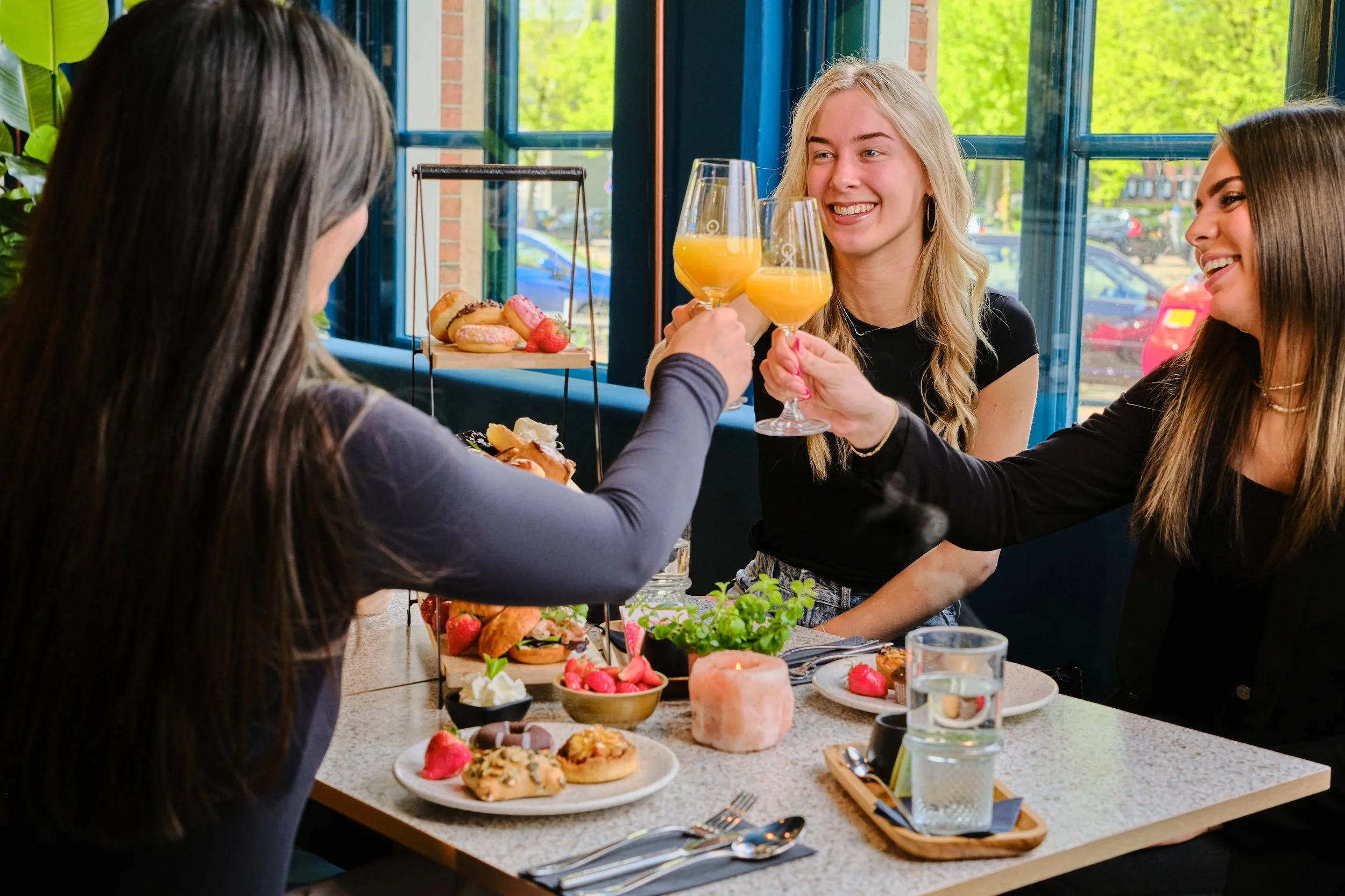Vrouw die toast met sinaasappelsap tijdens een brunch met drie vrouwen, met een etagère met gebak en fruit op de tafel.