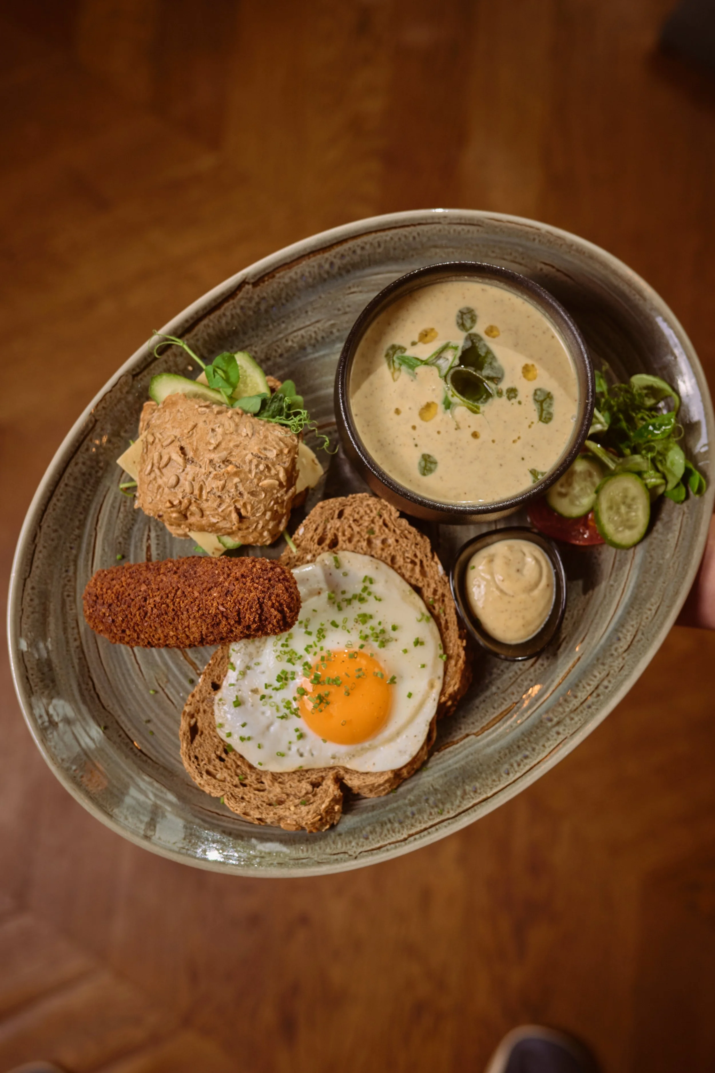 A plate of breakfast food with an over-easy egg on whole wheat toast, a breaded item, sliced cucumbers and cherry tomatoes, and a small bowl of creamy sauce, with a cup of creamy soup garnished with herbs.
