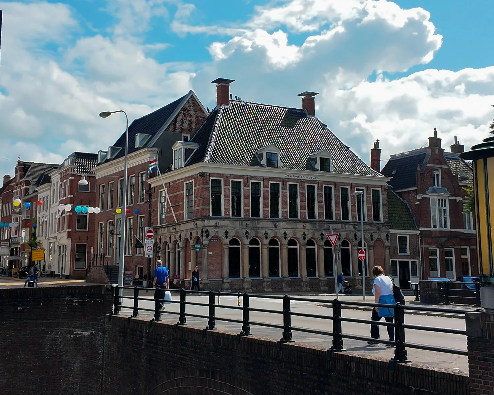 A street scene with historic brick buildings, some with European architectural features, under a partly cloudy sky. People are walking and riding bikes along the sidewalk, with colorful string lights and a street lamp visible.