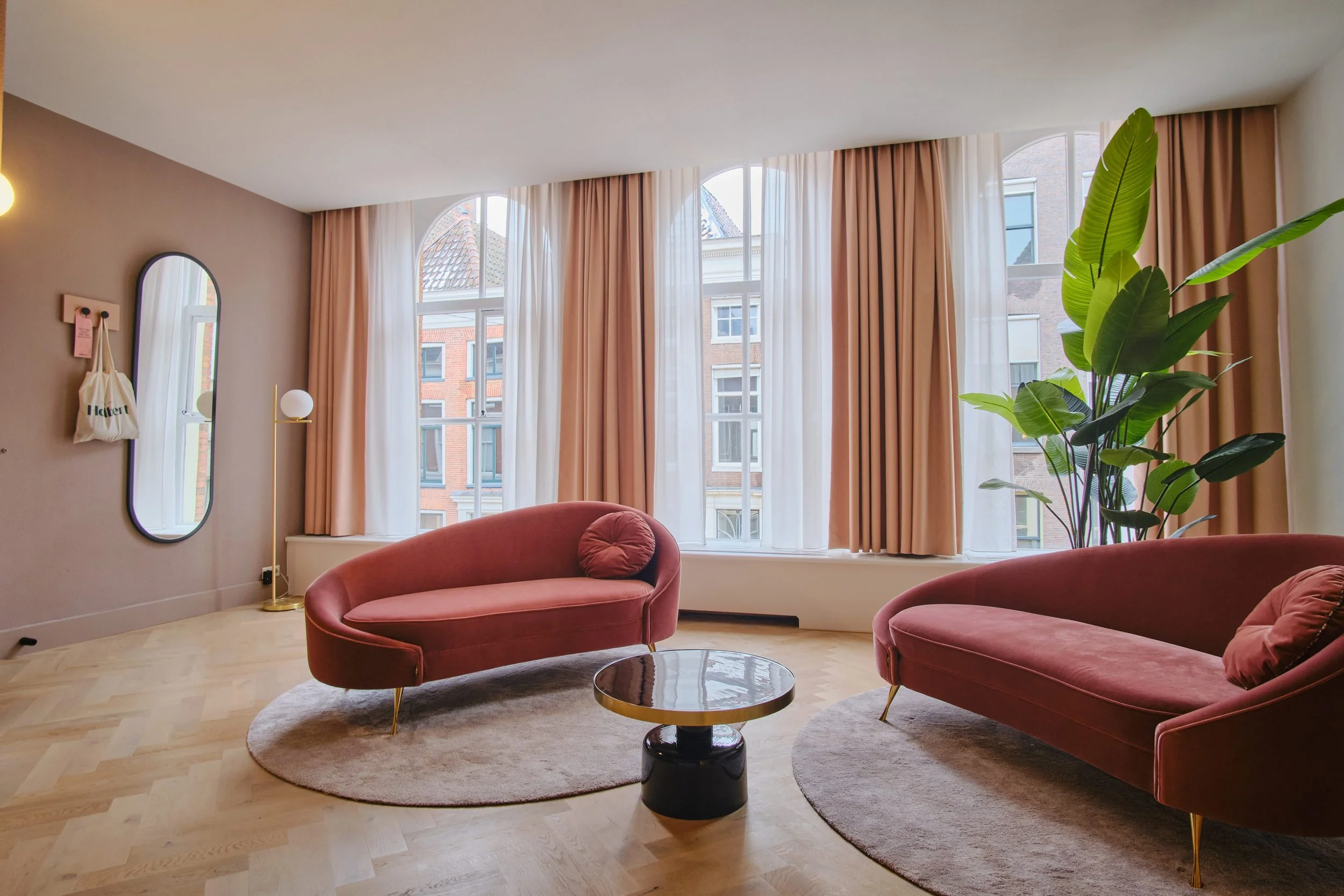 Living room with large windows, pink velvet sofas, a small round mirror, a tall floor lamp, a large green plant, a circular beige rug, a round coffee table, and beige curtains.