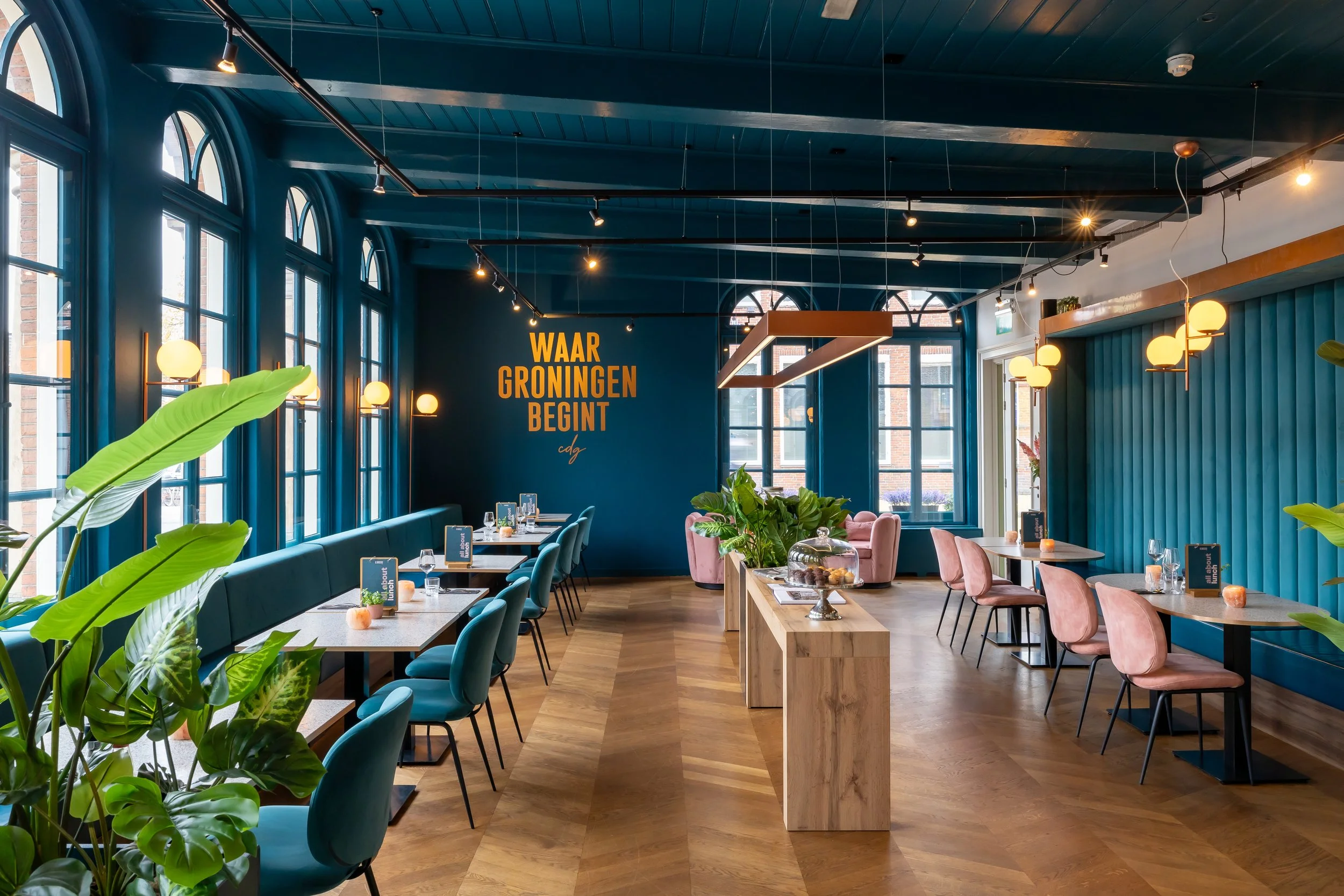 Interior of a modern restaurant with blue walls, large arched windows, wooden floors, and contemporary lighting fixtures. There are dining tables with chairs, a central wooden table with snacks, and a large sign on the wall that says 'WAAR GRONINGEN BEGINT' in yellow text.