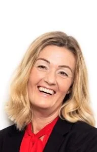 A woman with blonde hair smiling, wearing a black blazer and a red blouse against a white background.