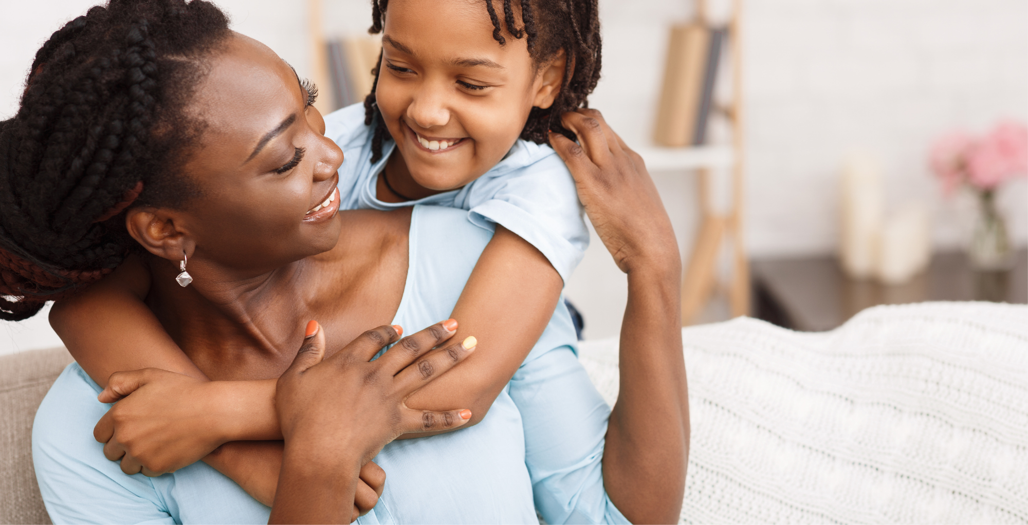 A woman and a young girl embracing and smiling at each other in a cozy, well-lit room with a blurred background featuring a bookshelf and a vase of pink flowers.