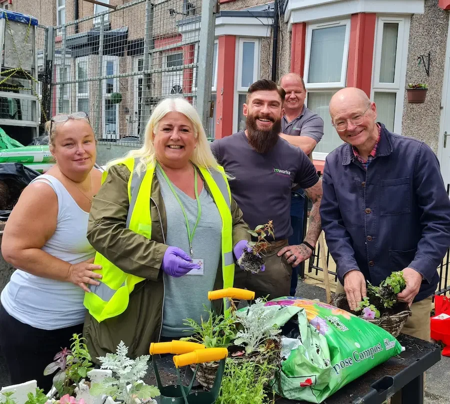 Group of five people smiling outdoors, planting flowers in a community garden, with gardening tools and plants on a table in front of them.