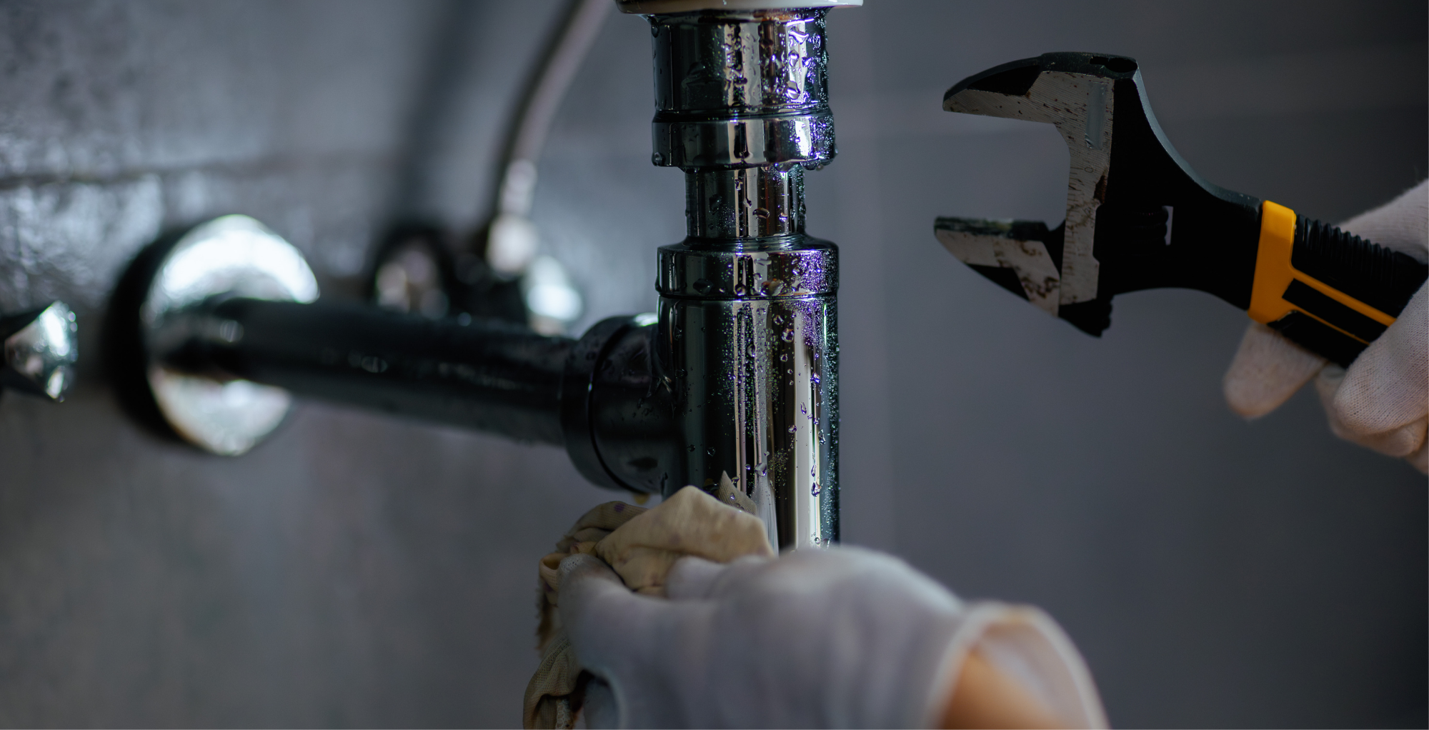 A person working on plumbing, tightening a pipe with a wrench, wearing gloves, with water droplets on the pipe.