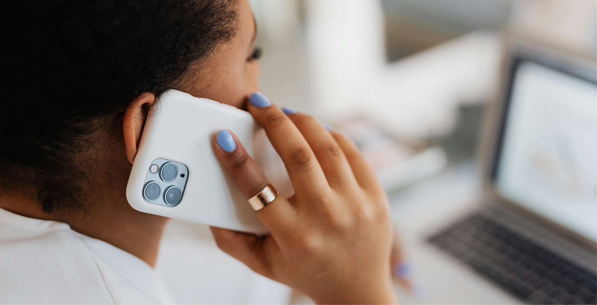 A woman with dark hair and painted nails is holding a white smartphone to her ear while sitting at a desk with a laptop open in front of her.