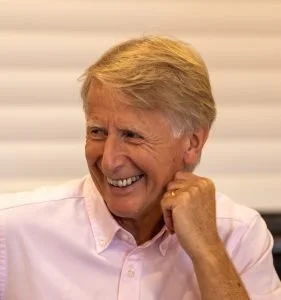 A smiling man with blonde hair wearing a light pink shirt, sitting indoors.