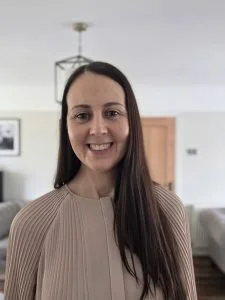 A woman with long dark hair smiling indoors, wearing a beige top.
