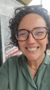 Close-up of a woman with curly dark hair, wearing glasses and a green shirt, smiling.