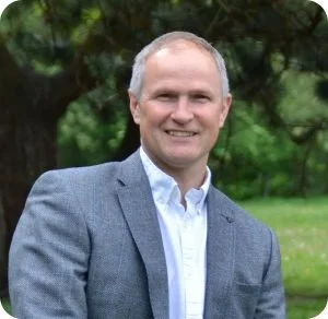 A smiling man in a gray blazer and white shirt outdoors with green trees in the background.
