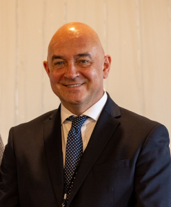 Headshot of a smiling man in a dark suit, white shirt, and patterned tie, standing indoors against a light background.
