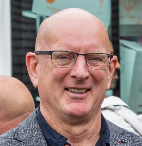 Headshot of a smiling man wearing glasses and a gray blazer, outdoors in an urban setting.