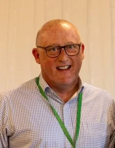 A smiling man with glasses wearing a checkered shirt and a green lanyard.