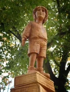 Wooden sculpture of a young boy with sunglasses, standing on a wooden pedestal outdoors among trees.