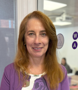 A woman with long red hair wearing a purple top, smiling in an office setting.