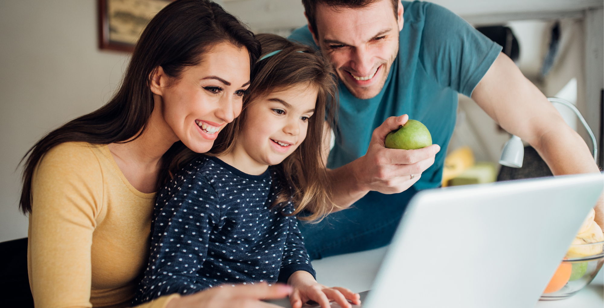 A family of three, a woman, a man, and a young girl, gathered around a laptop, smiling and looking at the screen together in a kitchen.