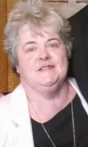 A woman with short  hair, wearing a black top and white jacket, smiling in a room with brown background.