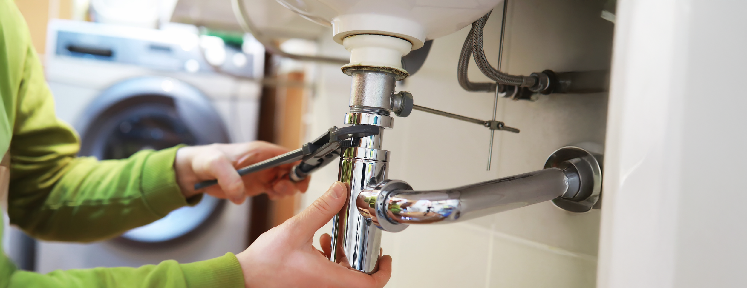Person wearing green shirt using pliers to repair or install a shiny chrome pipe under a sink in a laundry room.