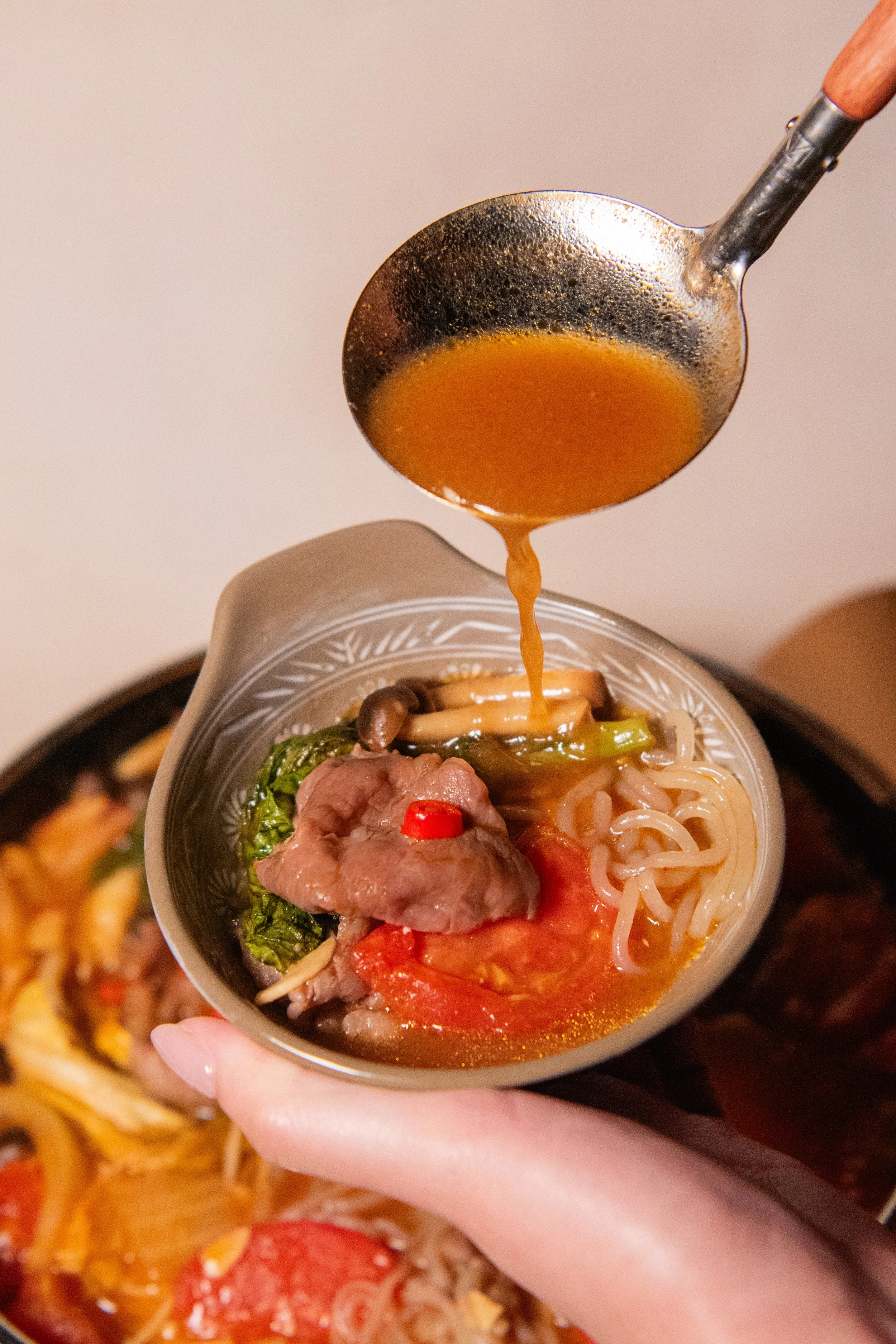 A hand holding a small bowl of ramen with broth, noodles, sliced meat, vegetables, and a red chili pepper, with a soup ladle pouring broth into the bowl.