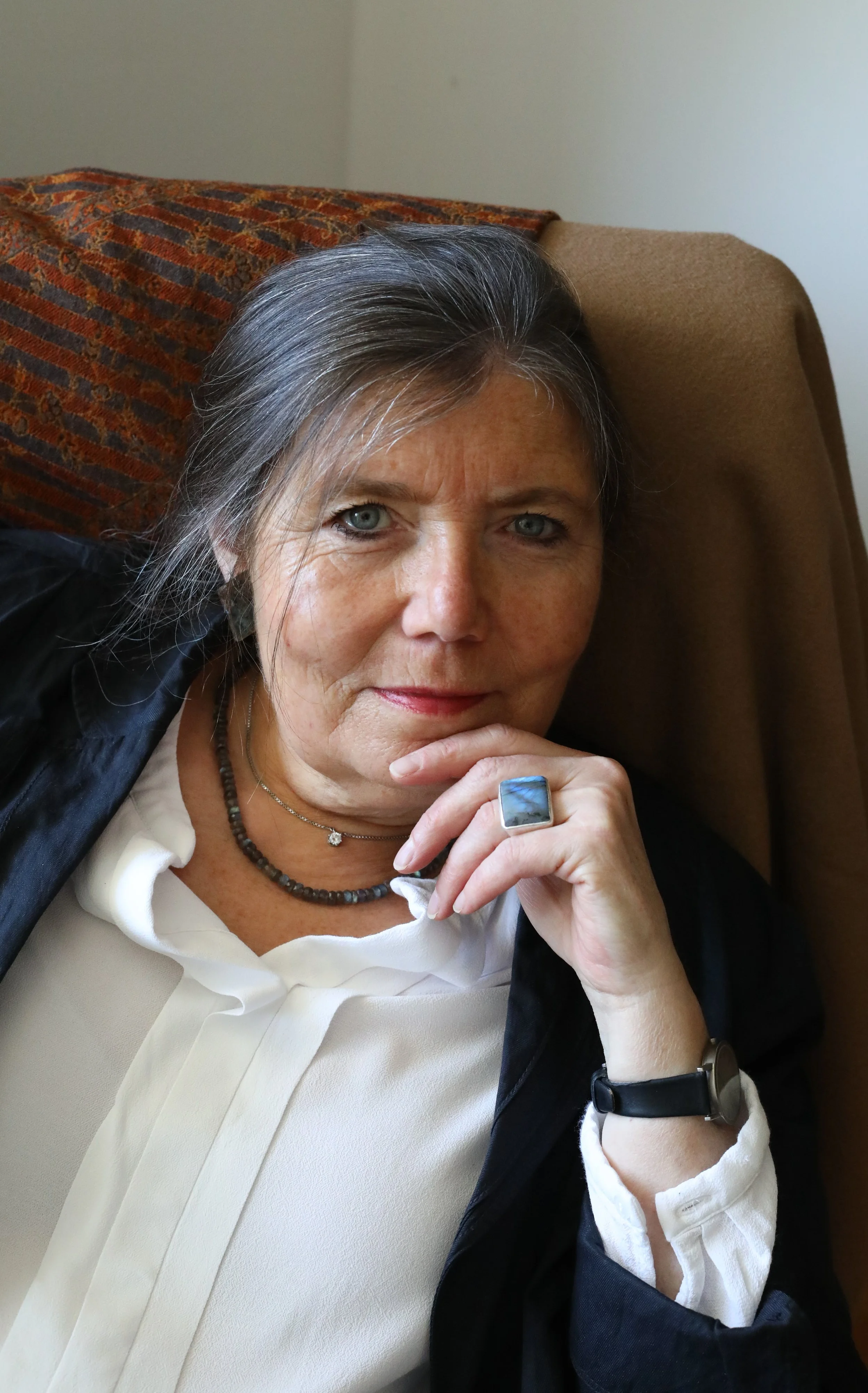 Eine ältere Frau mit grauen Haaren, blauen Augen und hellem Teint, die auf einem grauen Sofa liegt. Sie trägt eine weiße Bluse, eine dunkle Jacke, mehrere Schmuckstücke, darunter Ketten, Ringe und eine Armbanduhr, und schaut direkt in die Kamera. Im Hintergrund ist eine Wand und ein rotes, gemustertes Kissen.