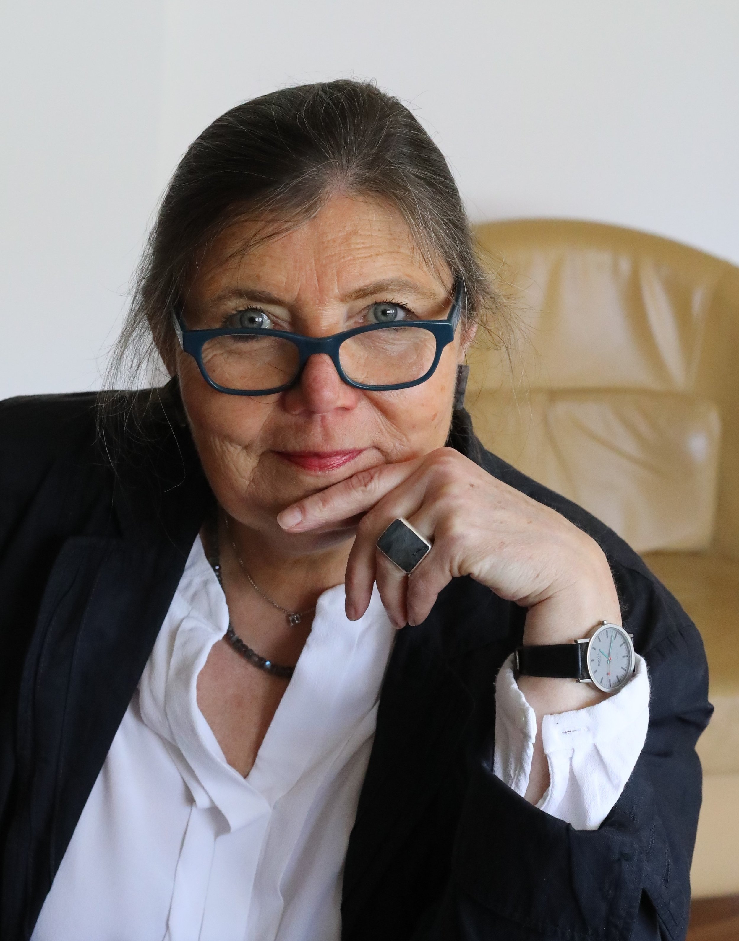 Eine ältere Frau mit dunklen Haaren, die eine Brille trägt, sitzt an einem Tisch in einem Raum mit hellem Hintergrund. Sie trägt eine weiße Bluse, einen schwarzen Blazer, eine Halskette und einen Ring am Finger. Sie wirkt nachdenklich und schaut durch die Brille, während sie die Hand vor den Mund hält. Auf ihrem linken Handguls ist eine Armbanduhr zu sehen.