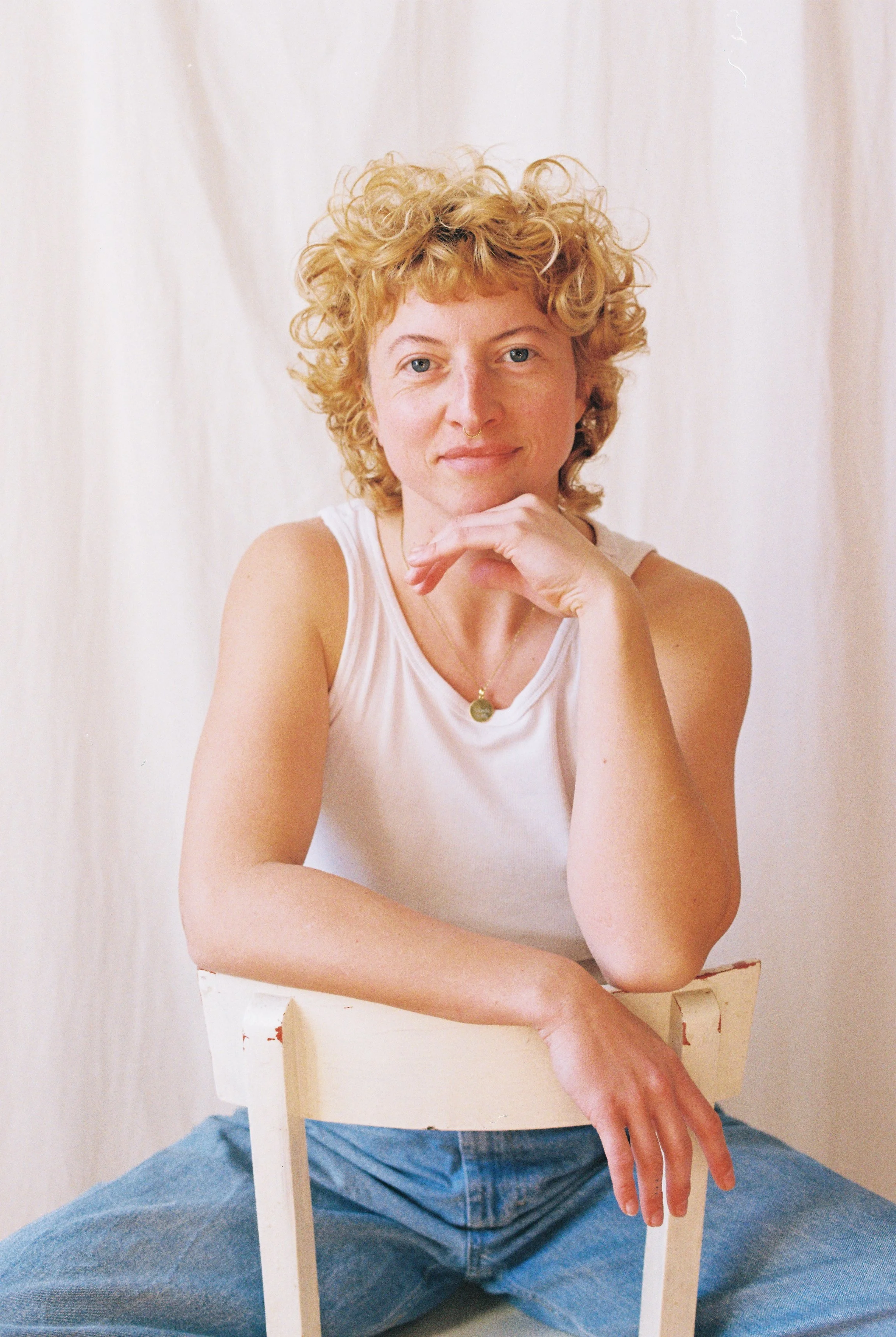 Junge Frau sitzt auf dem Boden, hält eine weiße Holzstühle und blickt lächelnd in die Kamera, trägt ein weißes Top und Jeans, im Hintergrund eine weiße Leinwand.