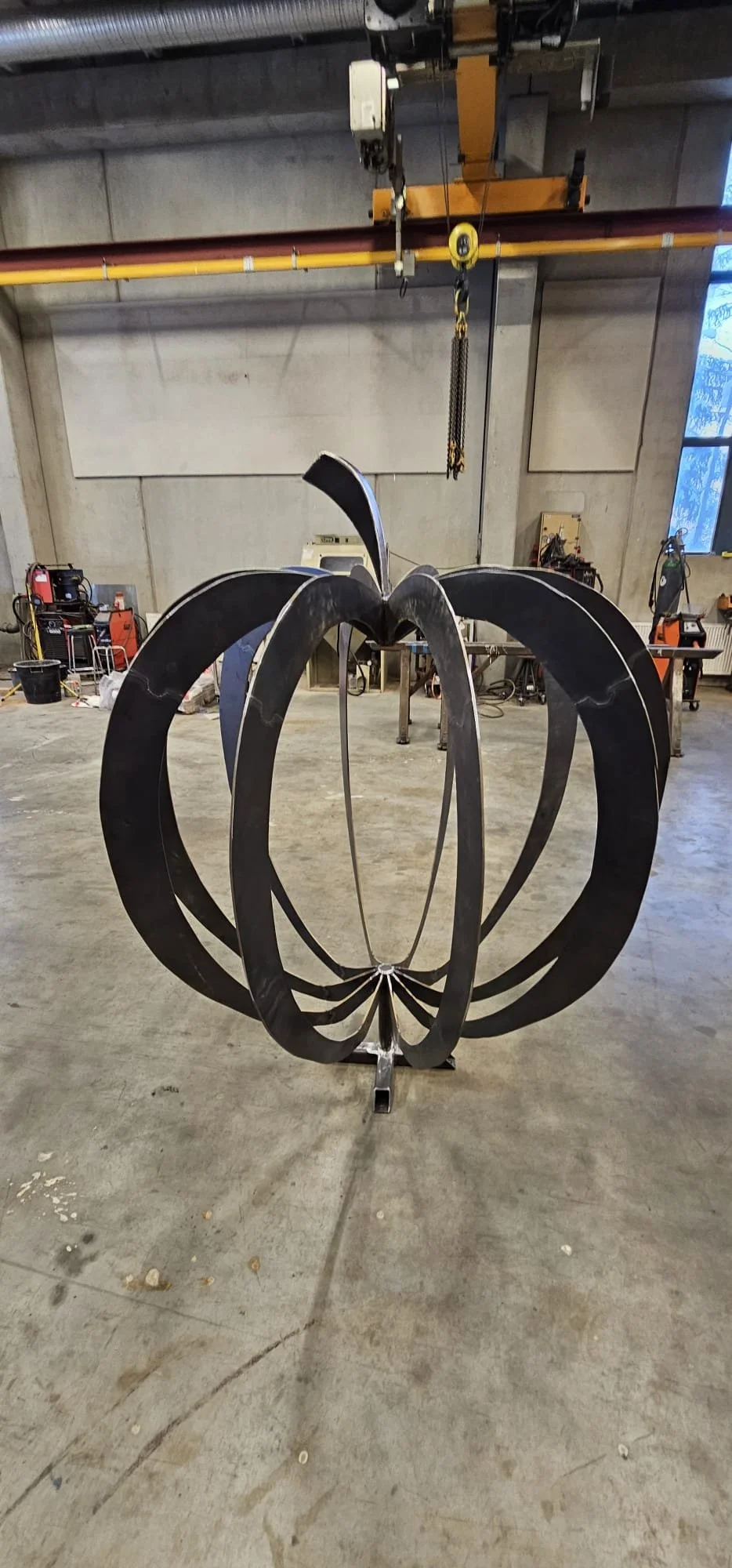 Large black metal sculpture resembling a stylized tulip or flower, suspended from a yellow crane hook inside an industrial workshop.