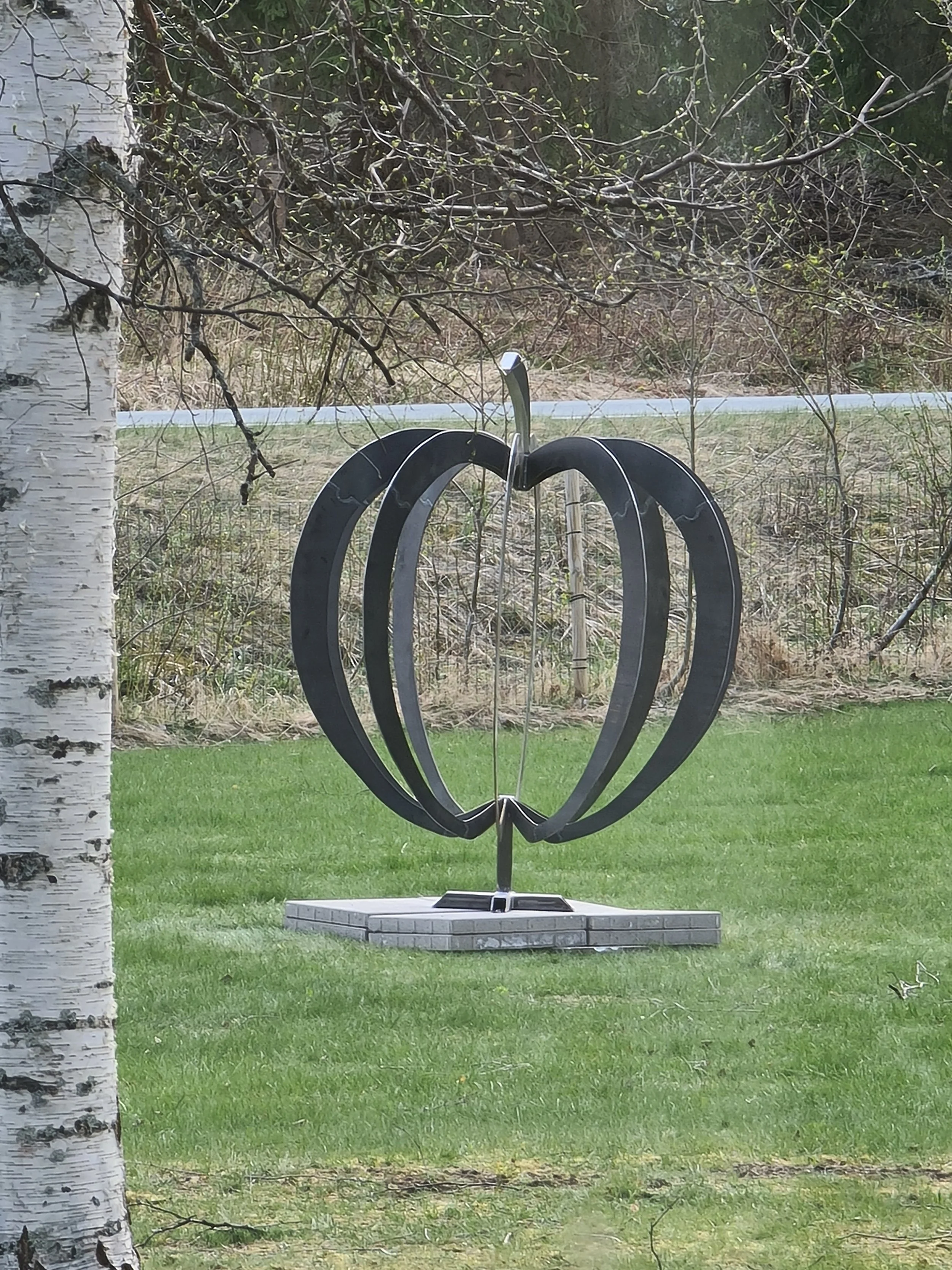A heart-shaped outdoor sculpture made of metal on a stone base, placed on a well-maintained grassy area with a tree trunk on the left and a wooded background.