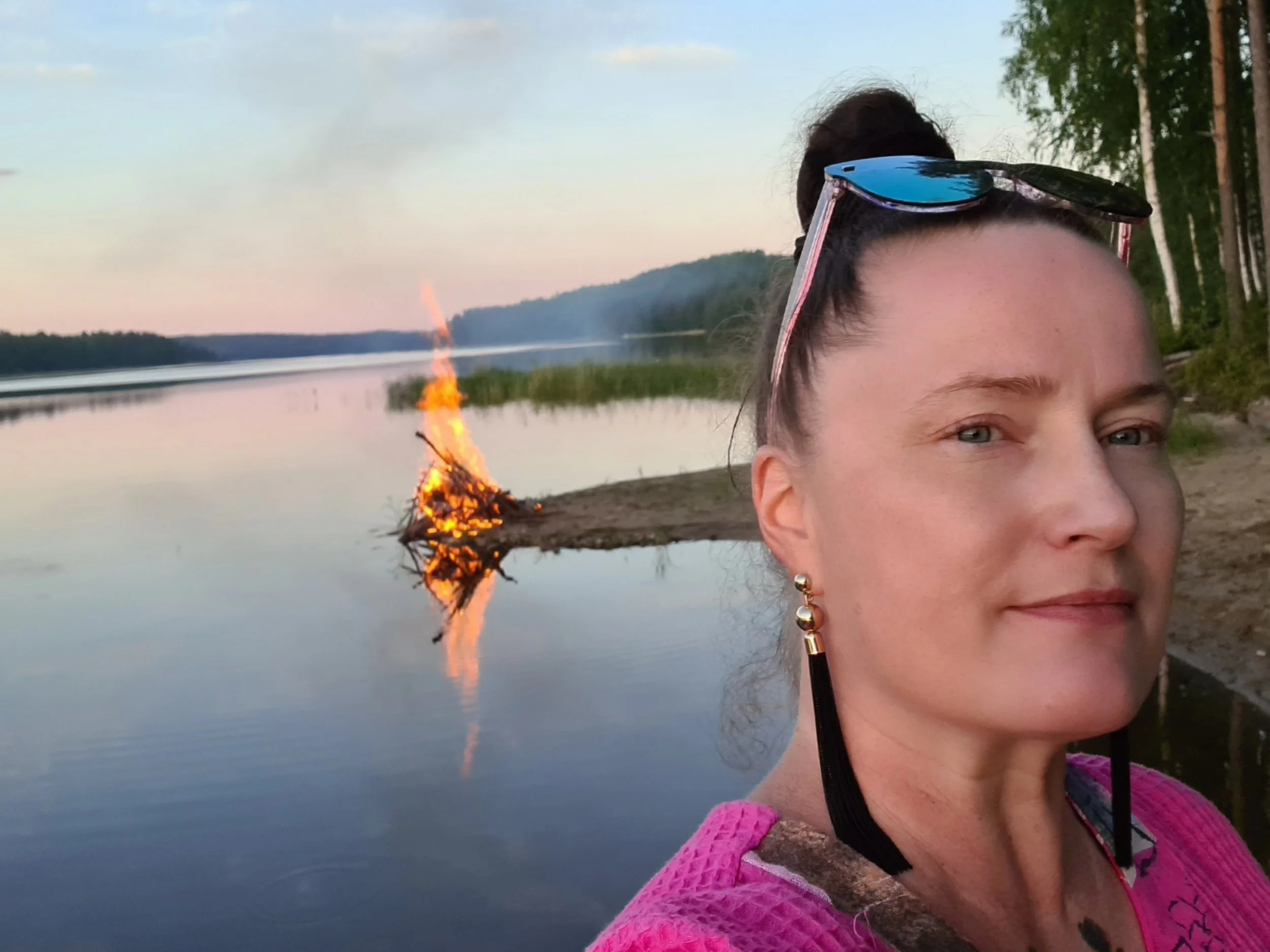 A woman with sunglasses on her head, earrings, and a pink sweater standing outdoors near a body of water with a small fire burning on the shoreline during sunset or twilight.