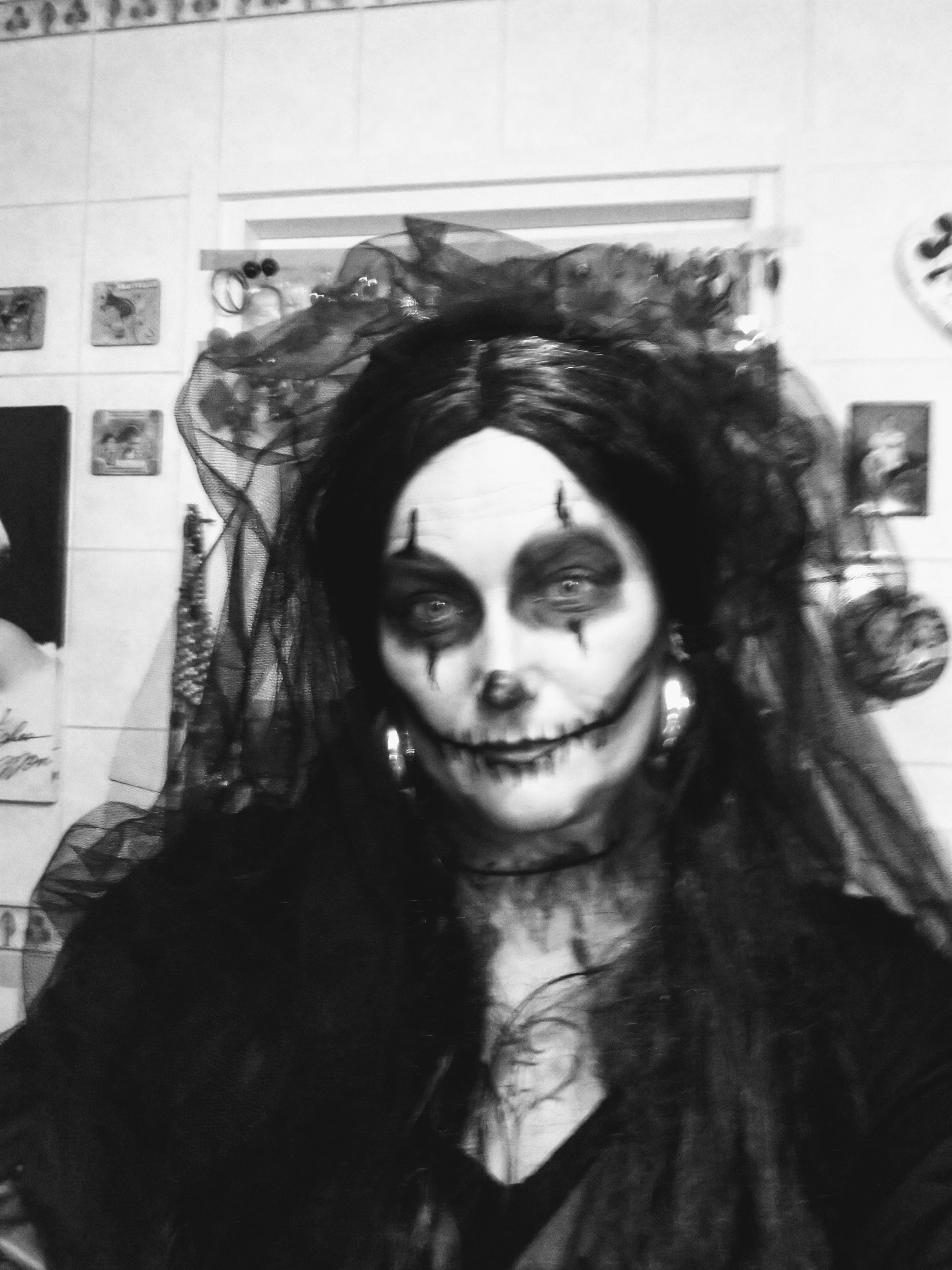 Black and white photo of a woman with painted skull face makeup, dark hair, and a lace veil or head covering. She is indoors with tiled walls and small framed pictures in the background.