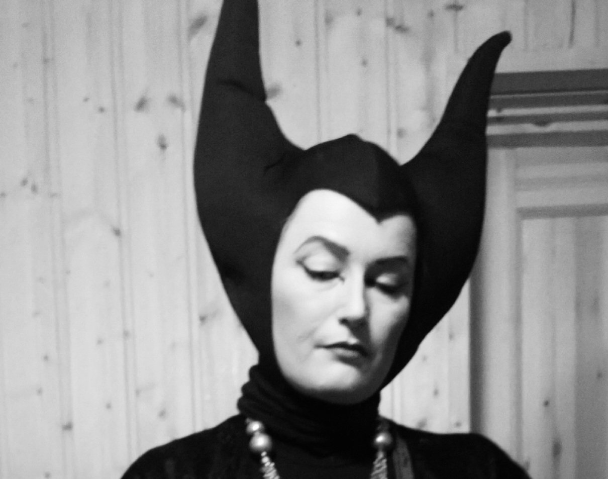 Black and white photo of a woman wearing a large, pointed headdress and a pearl necklace, standing in front of a wooden wall.