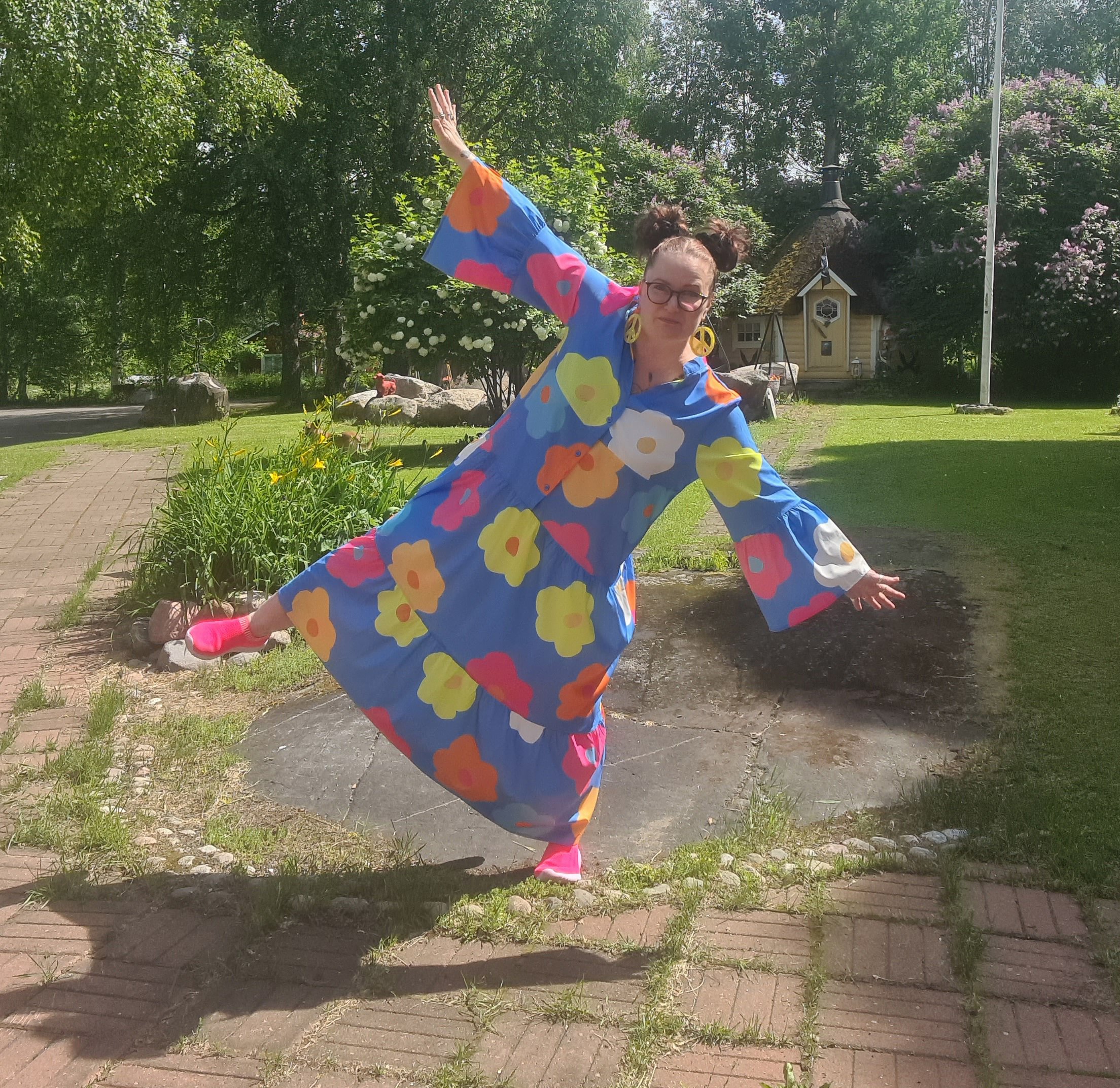 Woman in colorful floral dress striking a pose outdoors on a sunny day with trees, a small building, and a flagpole in the background.