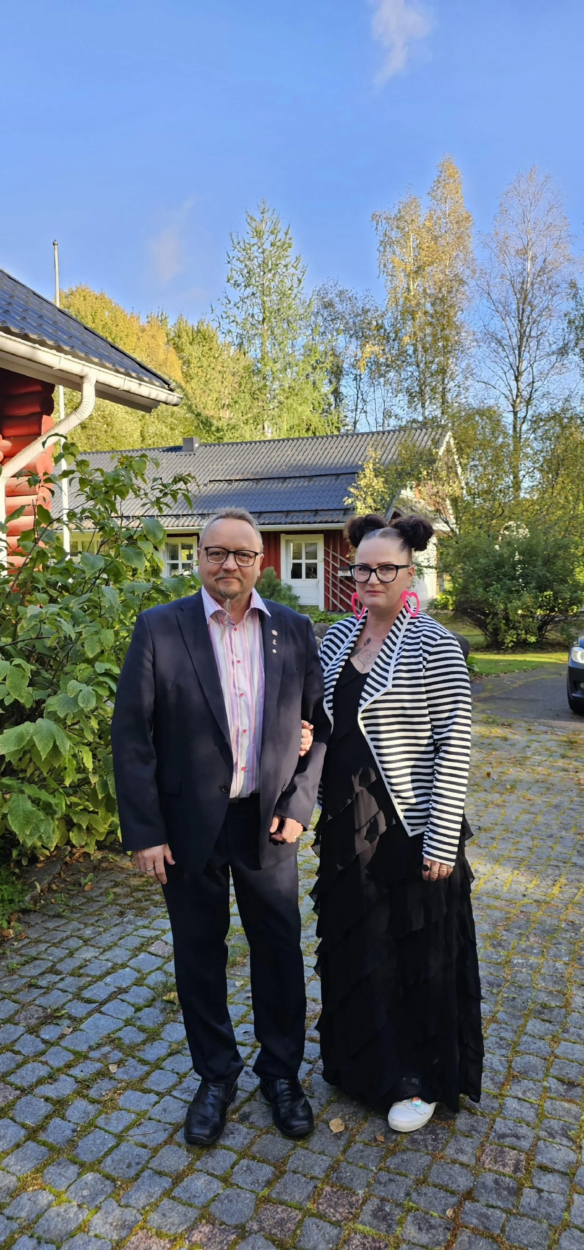 A man and a woman standing outdoors on a cobblestone path in front of green bushes and a red wooden house with a gray roof. The man is wearing a dark suit with a colorful striped shirt and glasses. The woman is wearing a black dress with a black and white striped blazer, large pink earrings, and sneakers, with her hair styled in two buns.