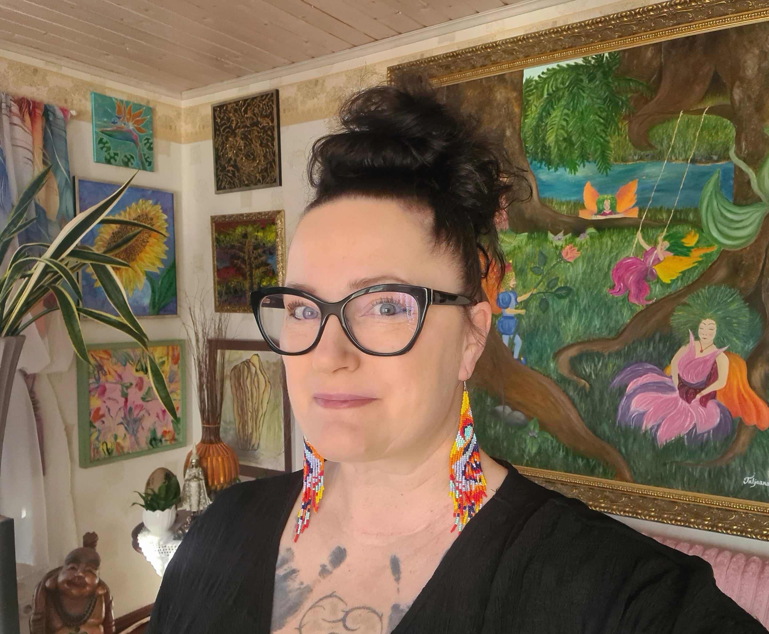 A woman with short dark hair, glasses, and colorful beaded jewelry is in an art gallery. The background displays vibrant paintings of nature and abstract art, and there are plants and decorative items visible.
