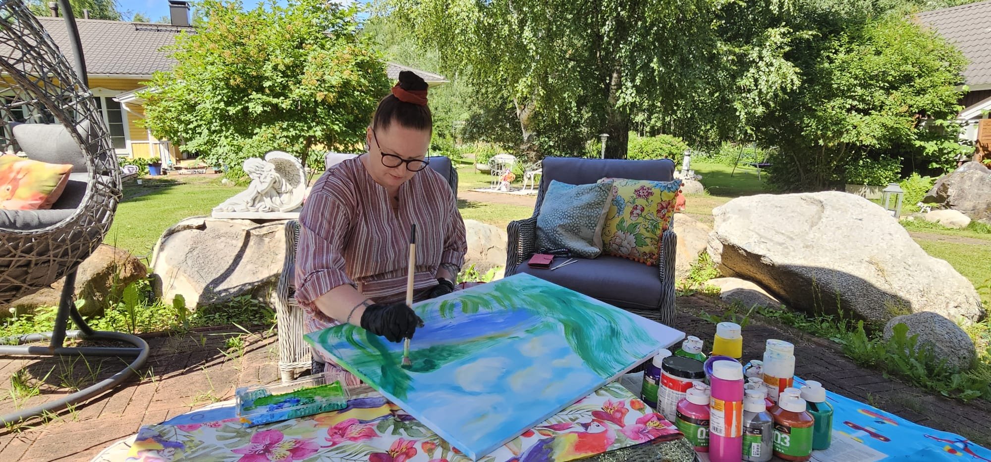 A woman with glasses, dark hair in a bun, and wearing a striped top, is painting outdoors on a sunny day. She is sitting on a wicker chair and working on a colorful landscape painting. There are multiple bottles of paint and brushes on the table in front of her. She is wearing black gloves and the background features trees, large rocks, and outdoor furniture.
